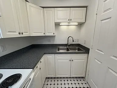 White kitchen cabinets with black countertop, stainless steel sink with a faucet, and a white oven in a small kitchen.