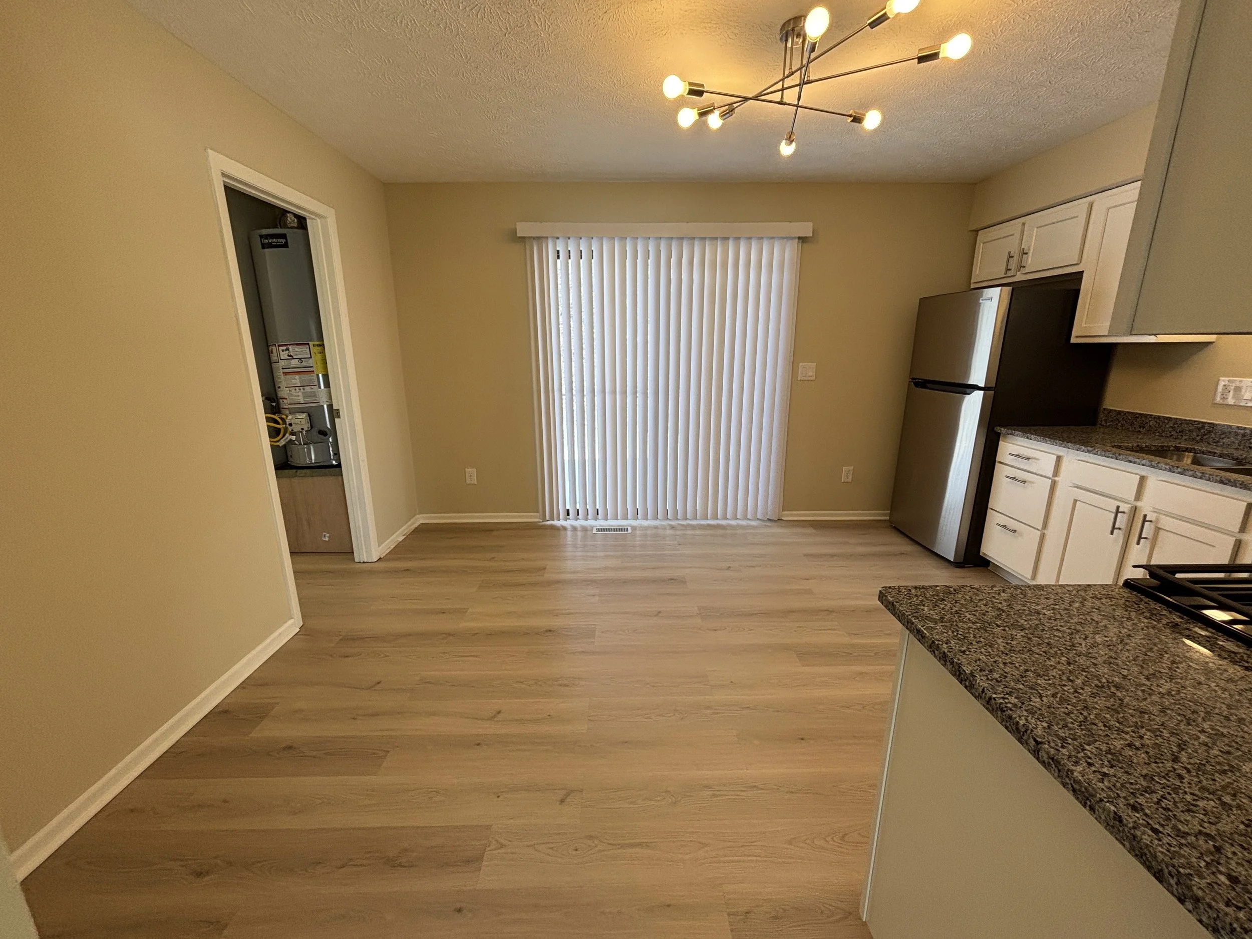 Empty kitchen and dining area with beige walls, light wood flooring, a sliding glass door with vertical blinds, modern ceiling light, stainless steel refrigerator, and white cabinets with granite countertops.