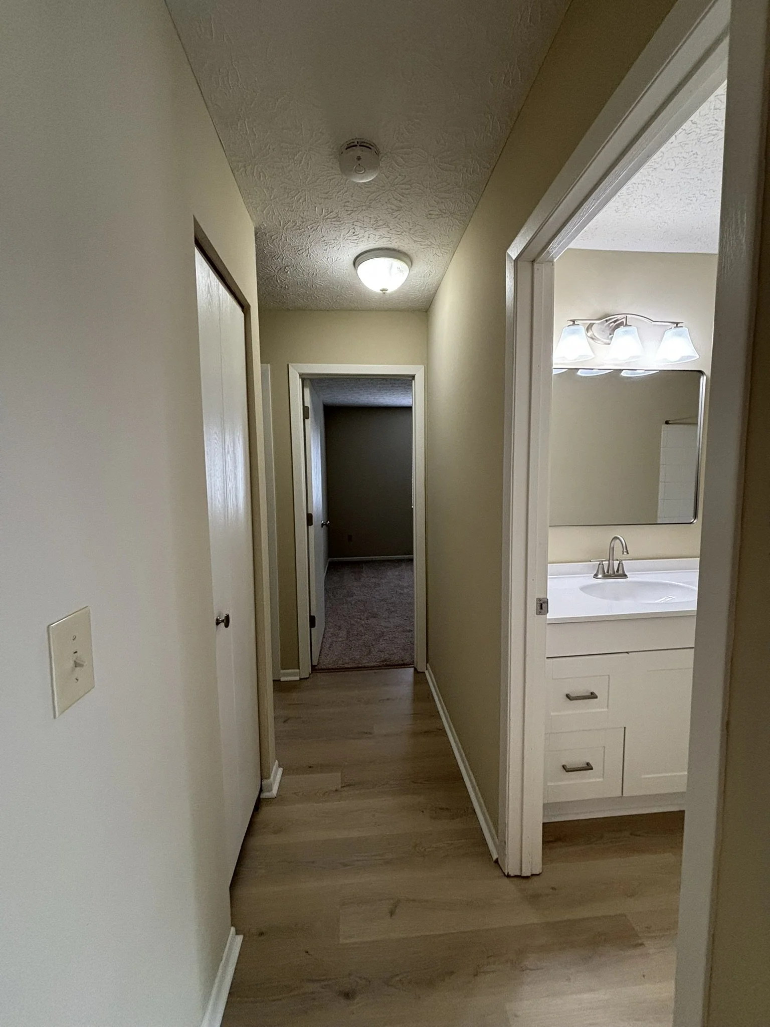 A hallway with beige walls, light wood flooring, and two ceiling lights leading to a darker room in the background. To the right, part of a bathroom with a white vanity, mirror, and light fixture is visible.