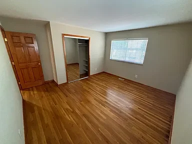 Empty room with hardwood floors, a window with blinds, a wooden closet door, and an open doorway leading to another room.