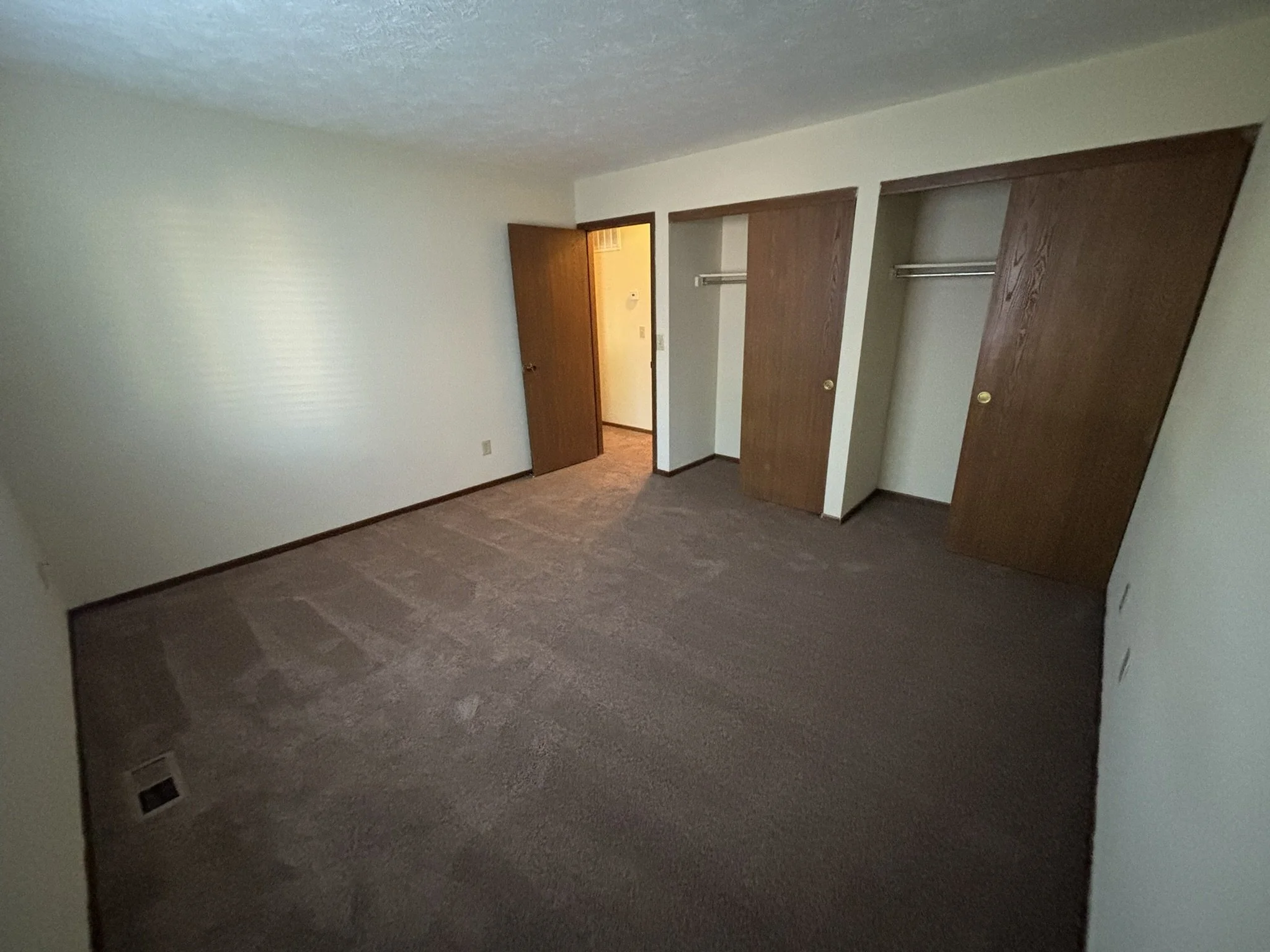 Empty bedroom with beige walls, brown carpet, and two wooden closet doors.