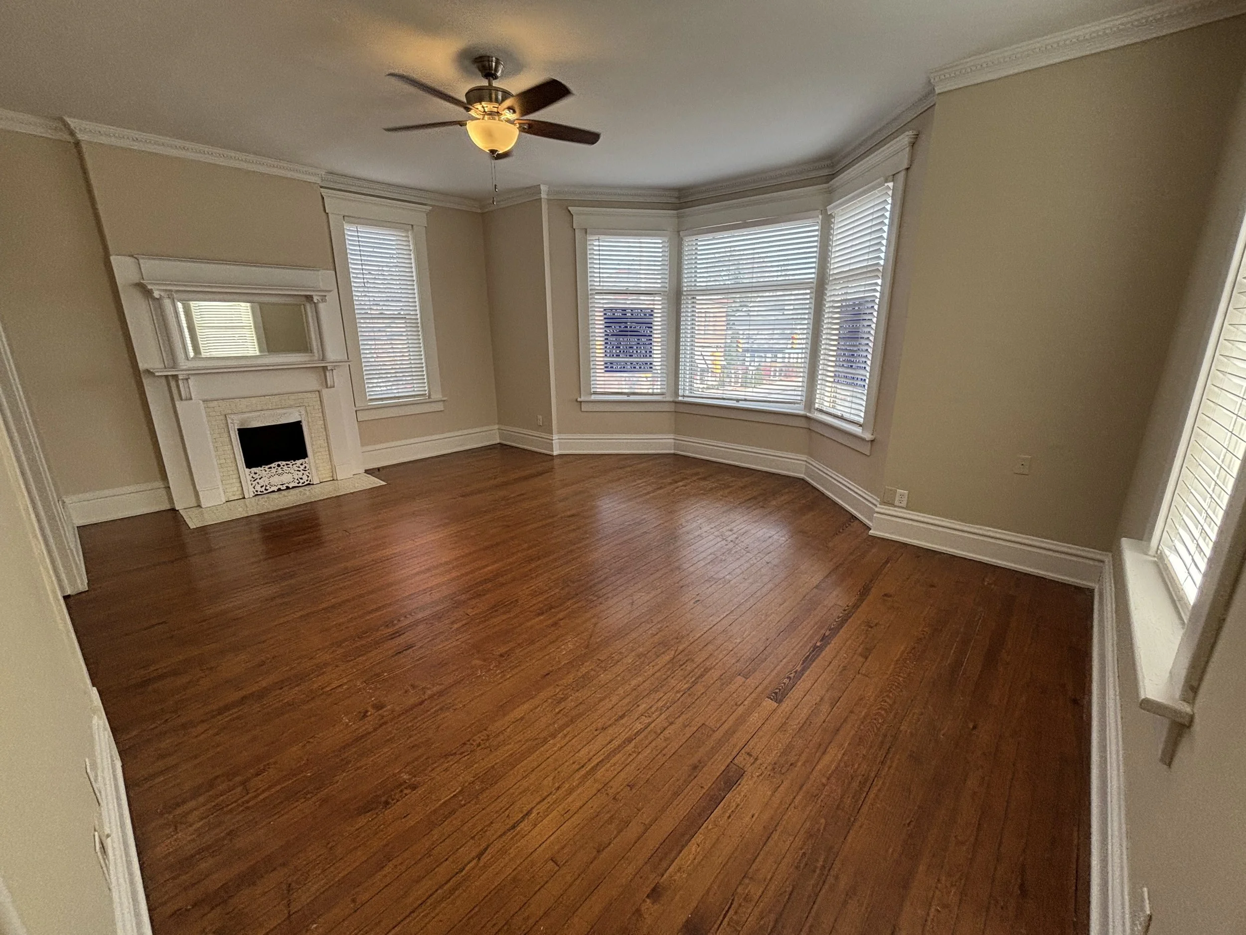 Empty living room with hardwood floors, bay windows with blinds, a ceiling fan, and a decorative fireplace with a mirror above it.