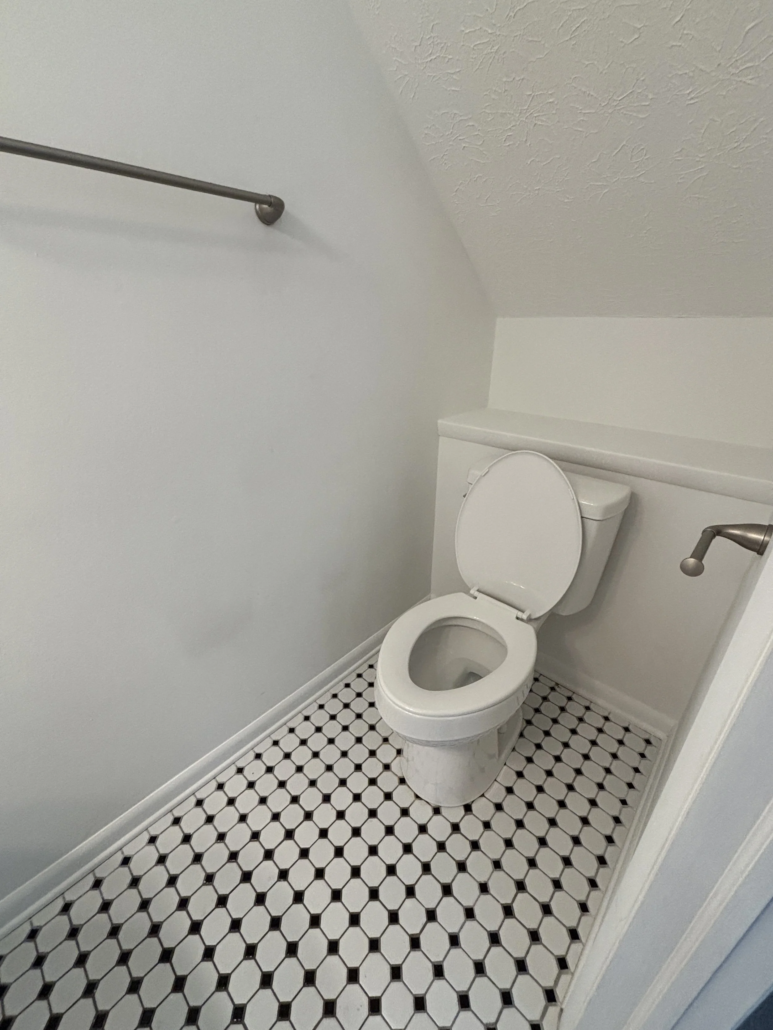 A small bathroom with a white toilet, black and white hexagonal tile flooring, a towel bar on the wall, and a textured ceiling.