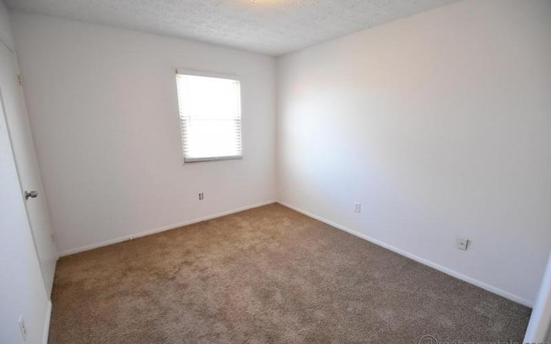 Empty room with white walls, a window with blinds on the back wall, a closed door on the left, and carpeted beige floor.