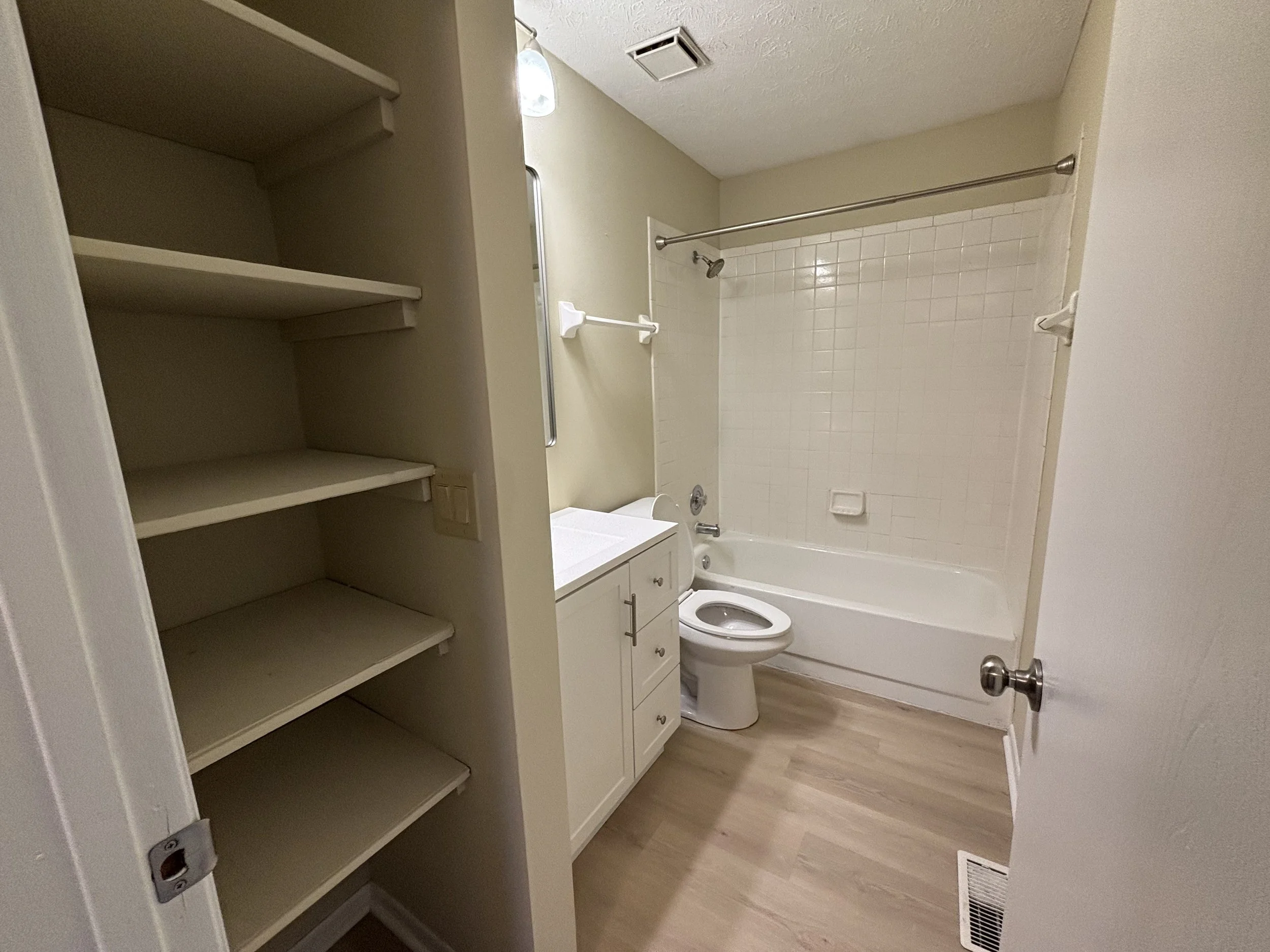 Small bathroom with a bathtub and shower, toilet, white vanity, and empty storage shelves.