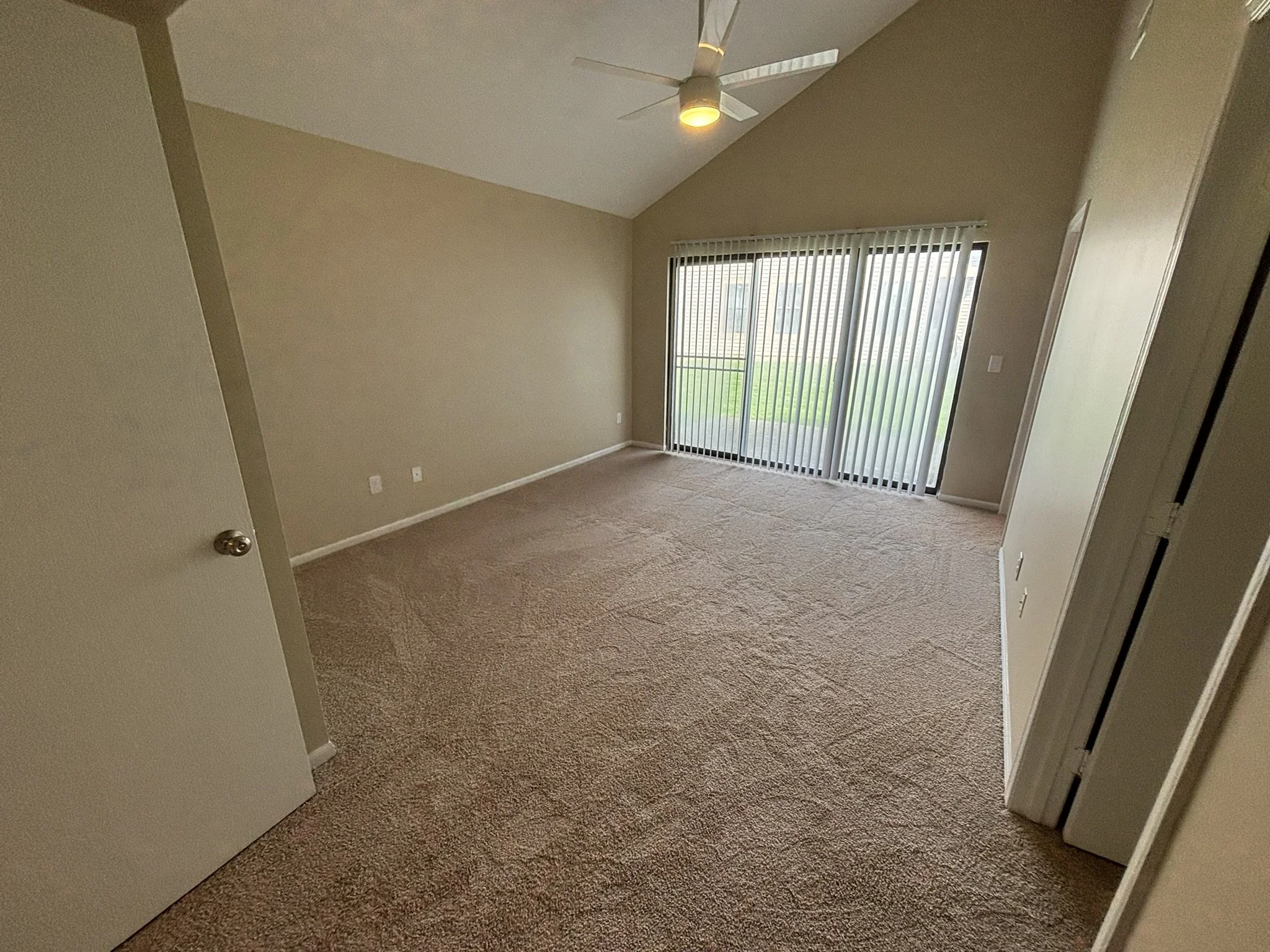 Empty living room with beige carpet, sliding glass door with vertical blinds, and ceiling fan with light.