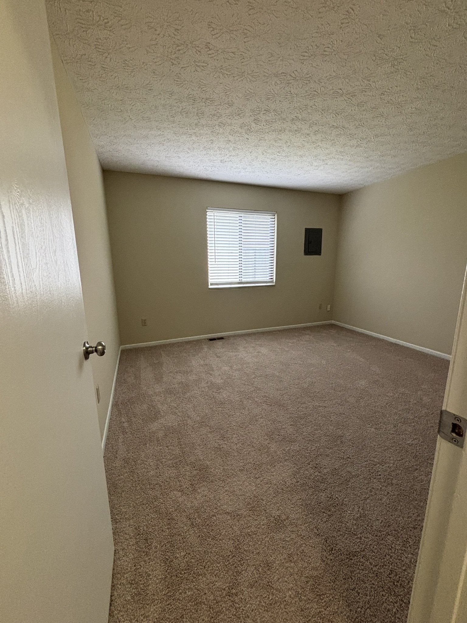 Empty room with beige carpet, beige walls, a window with blinds, and an air vent on the wall.