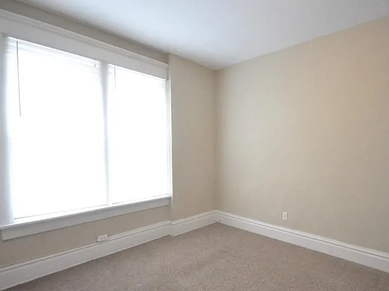 Empty room with beige walls, white baseboards, a carpeted floor, and a large window with blinds letting in natural light.