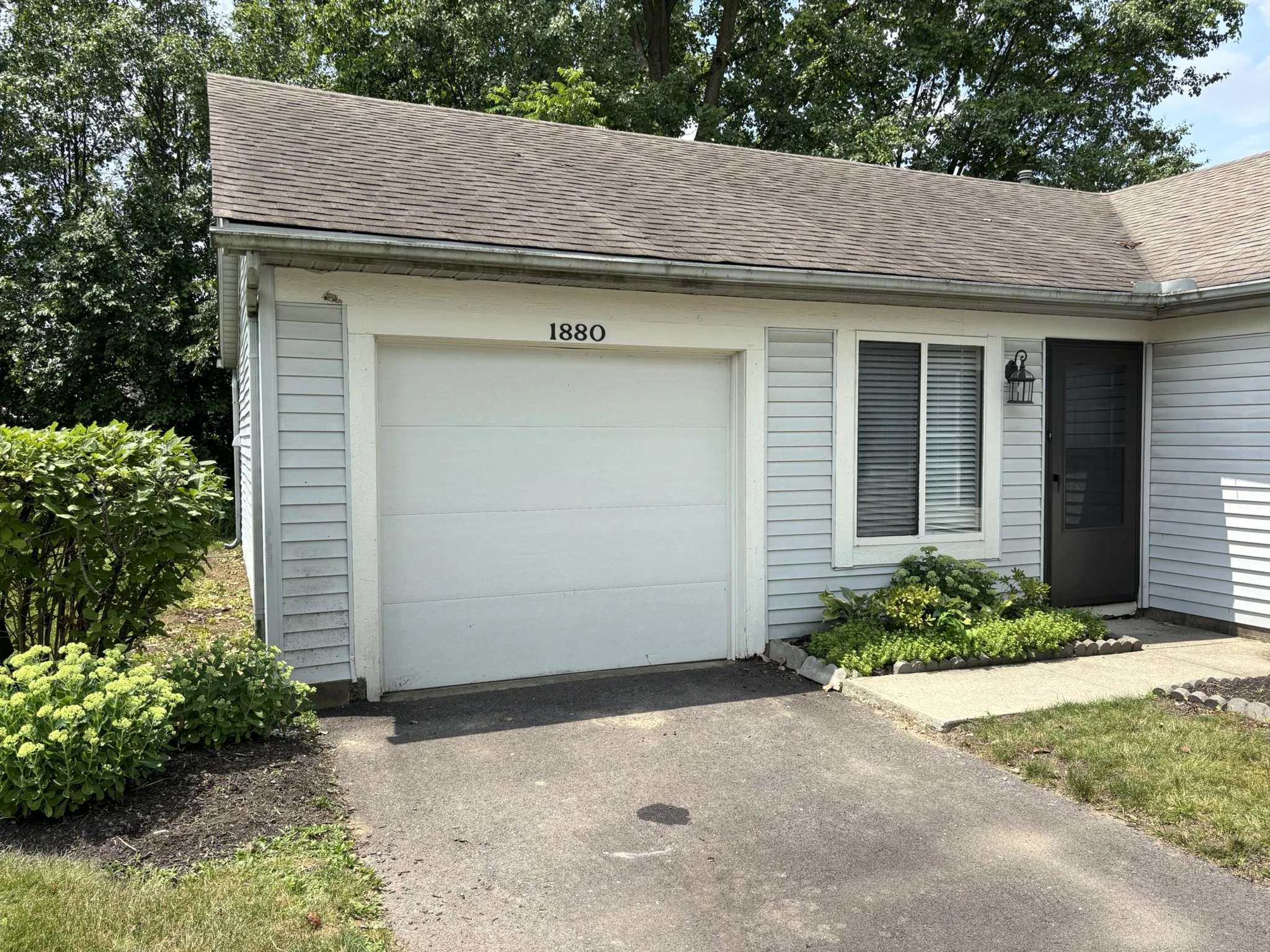 View of a house with gray siding, a white garage door numbered 1880, and a black front door. There are small flower beds with green plants in front of the house and a concrete path leading to the front door. A black light fixture is mounted beside th