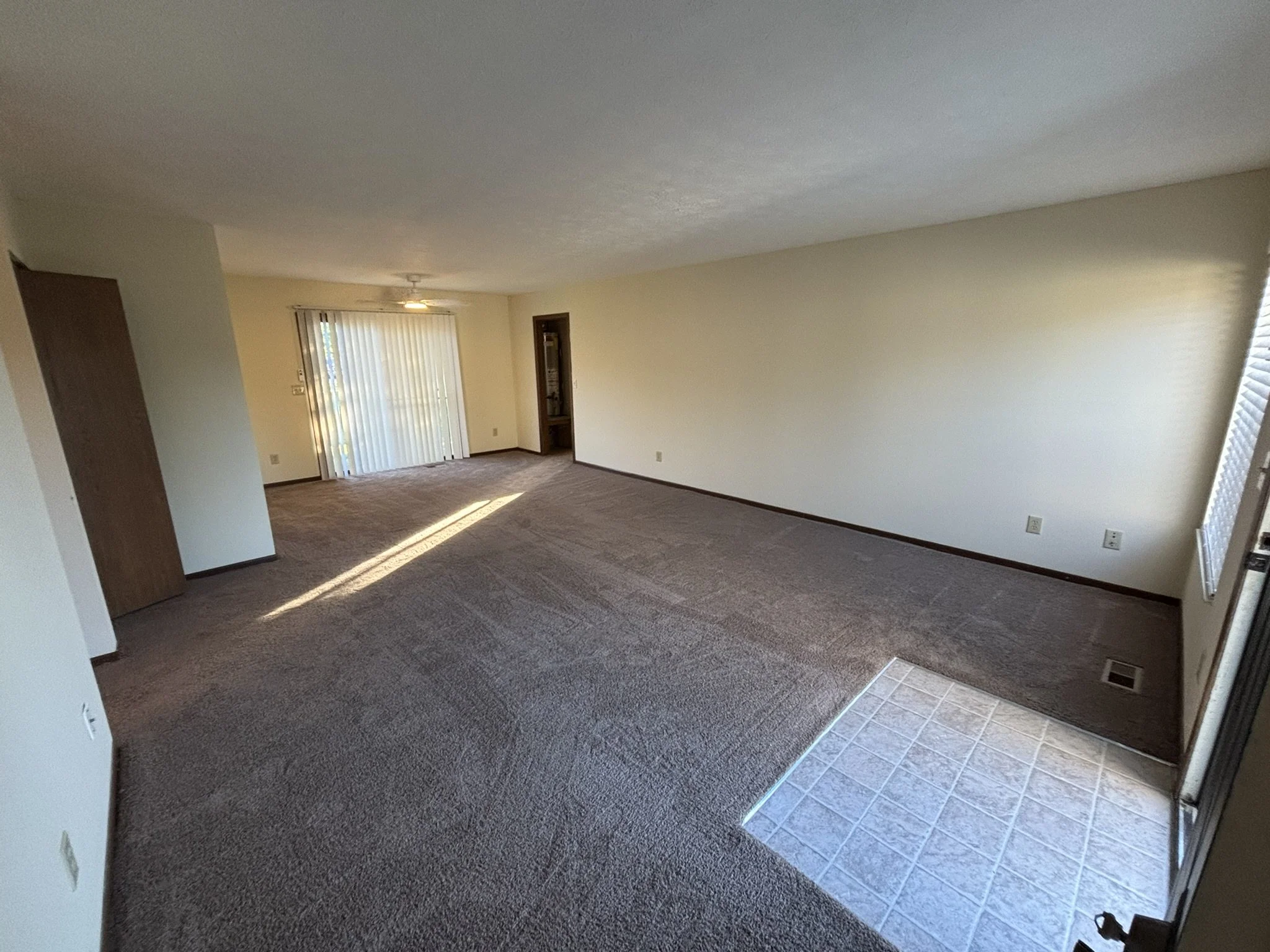Empty living room with beige carpet, white walls, sliding glass door with vertical blinds, a ceiling fan, and a tiled area near the doorway.