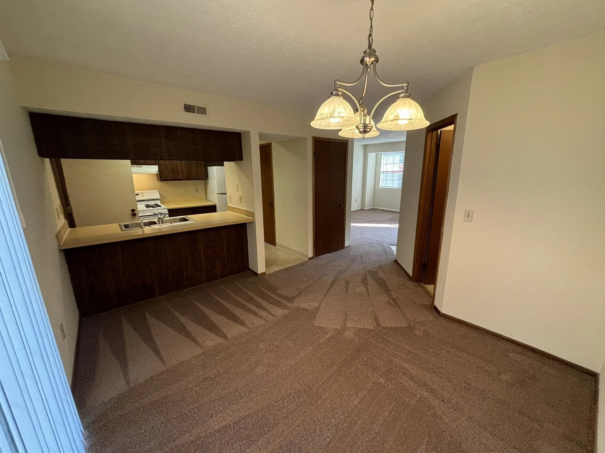 Empty dining area with a hanging light fixture, dark wood kitchen cabinets, and a view into a room with a window and carpeted floor.