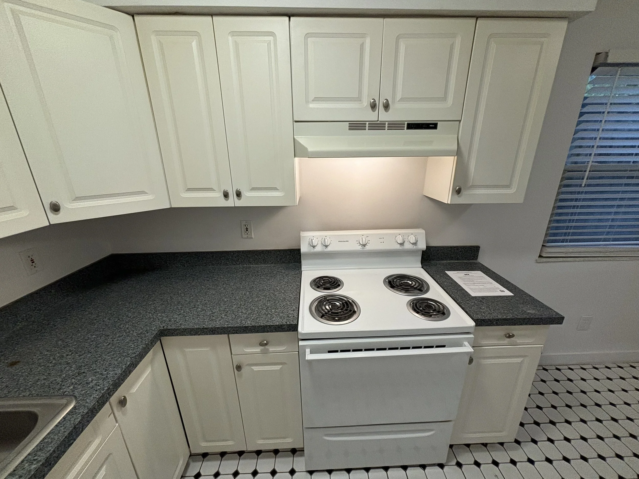 White kitchen stove with four coil burners and a control panel, surrounded by white cabinets and a black speckled countertop, with a window with blinds on the right.