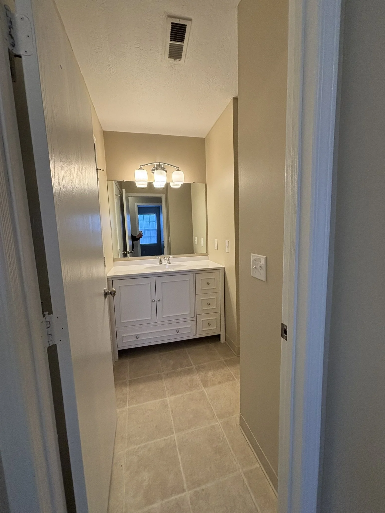 A small bathroom with light beige walls, a vanity with a white cabinet, a sink, and a large mirror above it. There are four ceiling lights above the mirror and a vent on the ceiling. The bathroom has beige tiled flooring.
