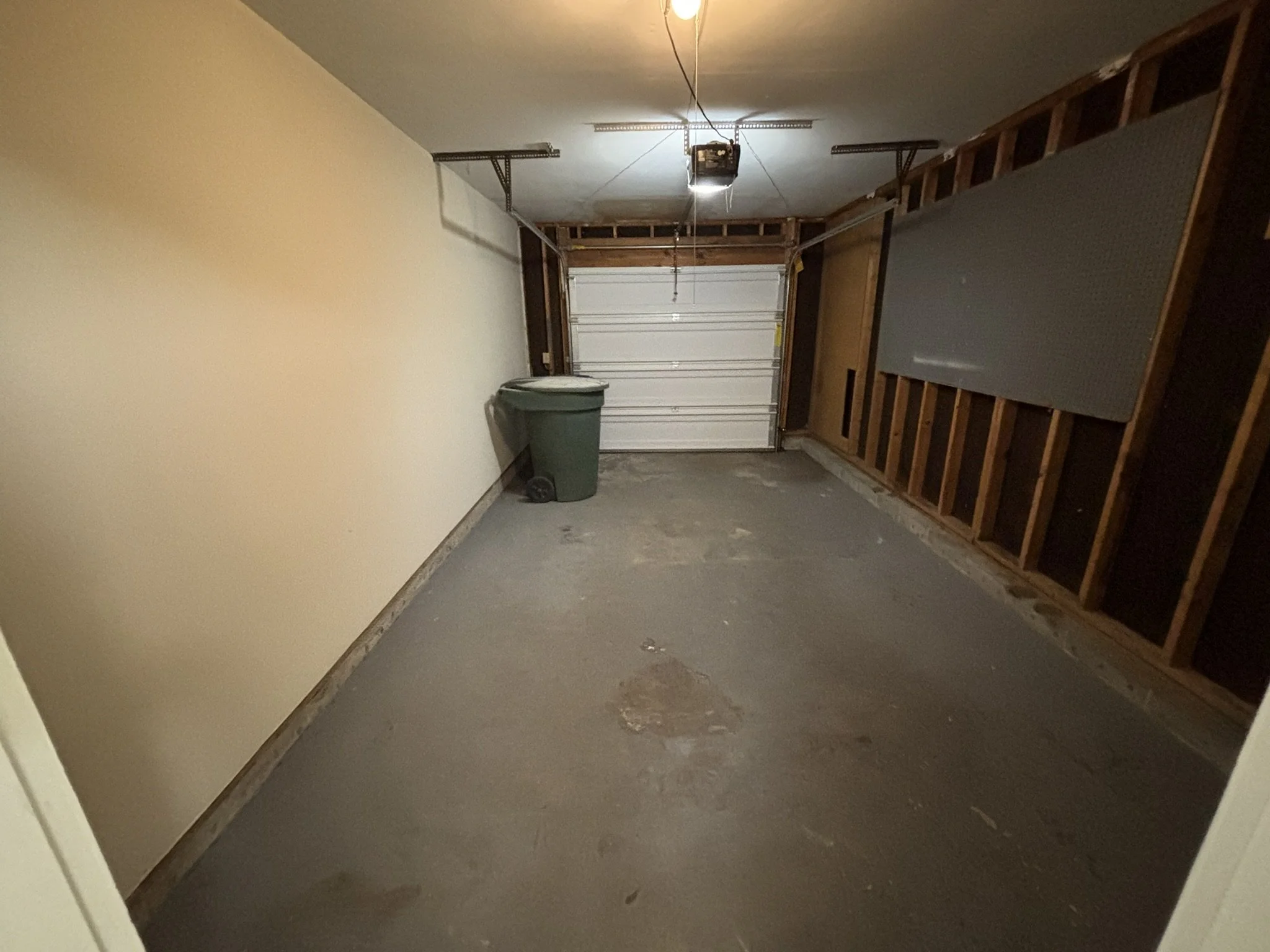 Empty garage with a green trash can, a closed white garage door, and exposed wooden framing on one wall.