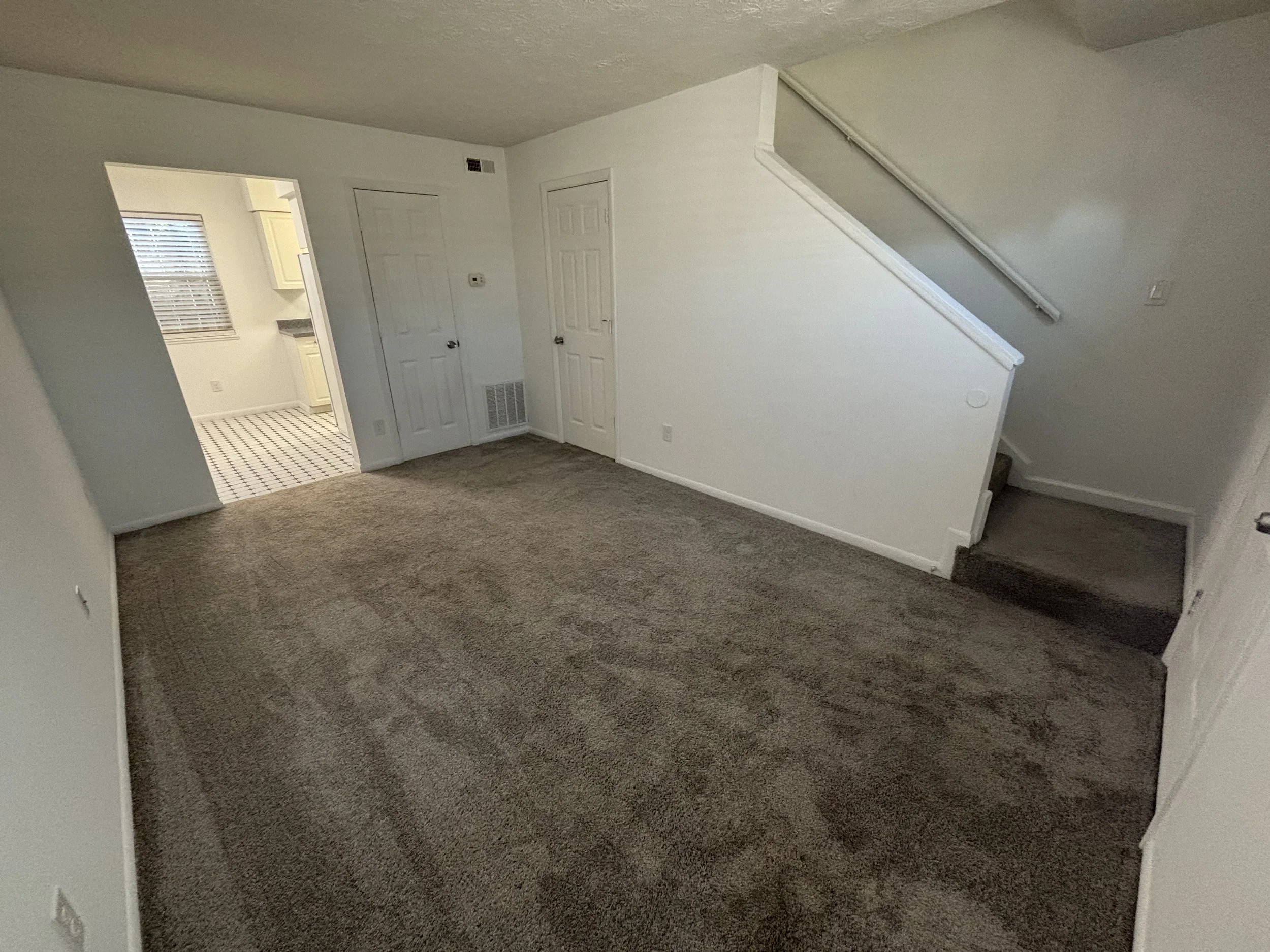 Empty living room with brown carpet, white walls, and staircase leading upstairs, with adjacent kitchen area visible through an open doorway.