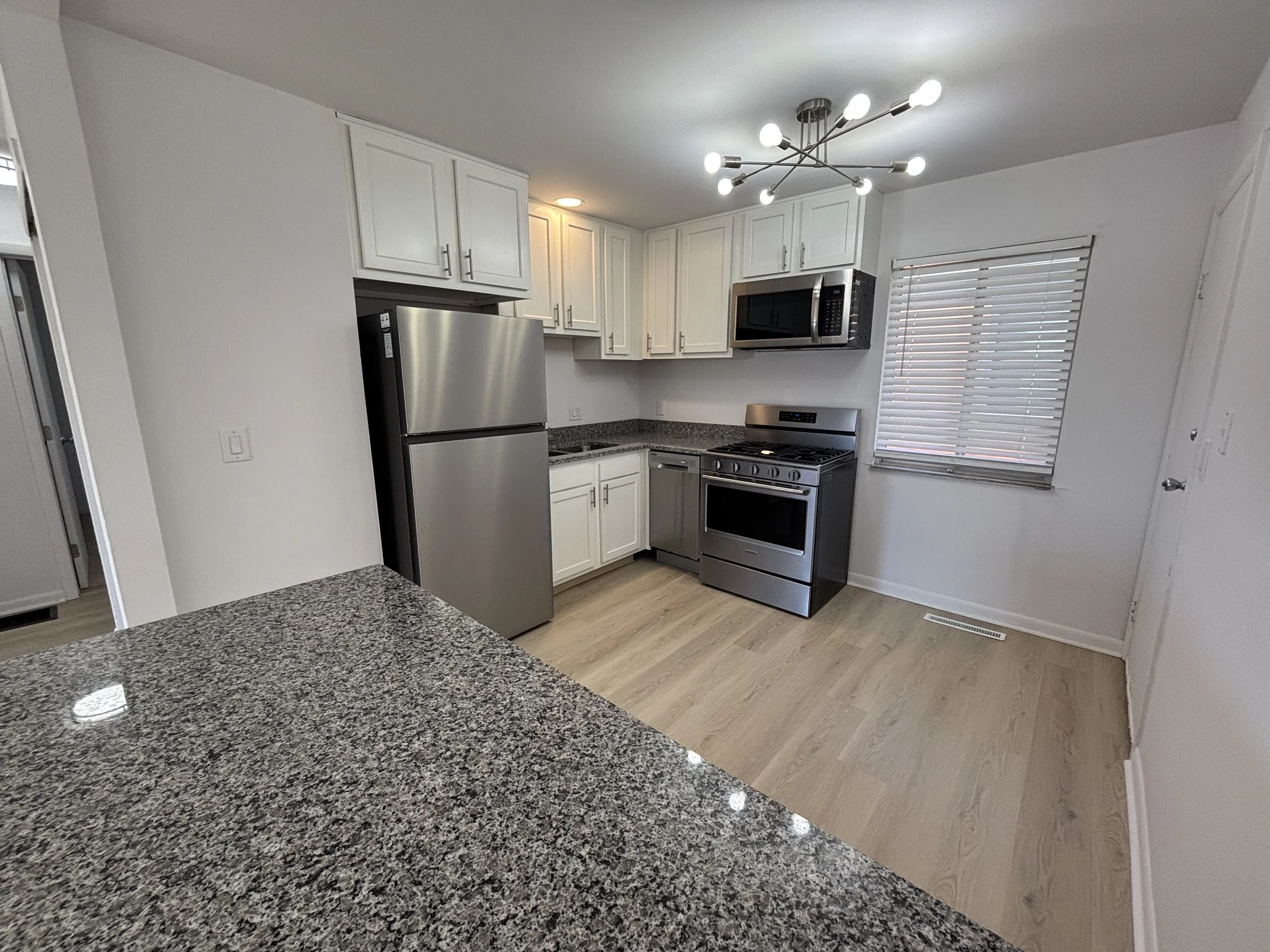 White kitchen with stainless steel refrigerator, stove, and microwave, granite countertops, white cabinets, and a window with blinds.