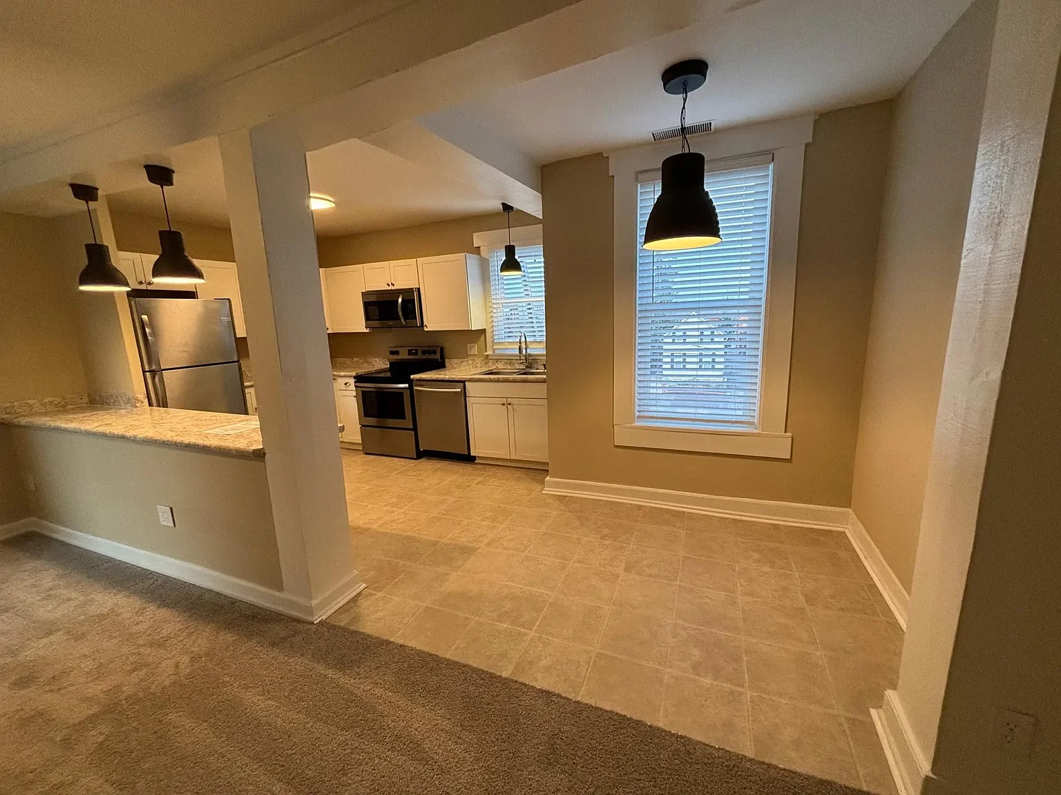 Empty dining area with two large windows and a hanging black pendant light, adjacent to a kitchen with beige cabinets, stainless steel appliances, and a granite countertop.
