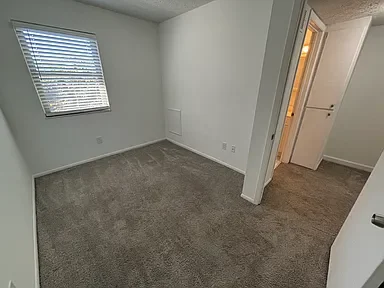 Empty room with beige carpet, white walls, a window with blinds, and an open doorway leading to another room.