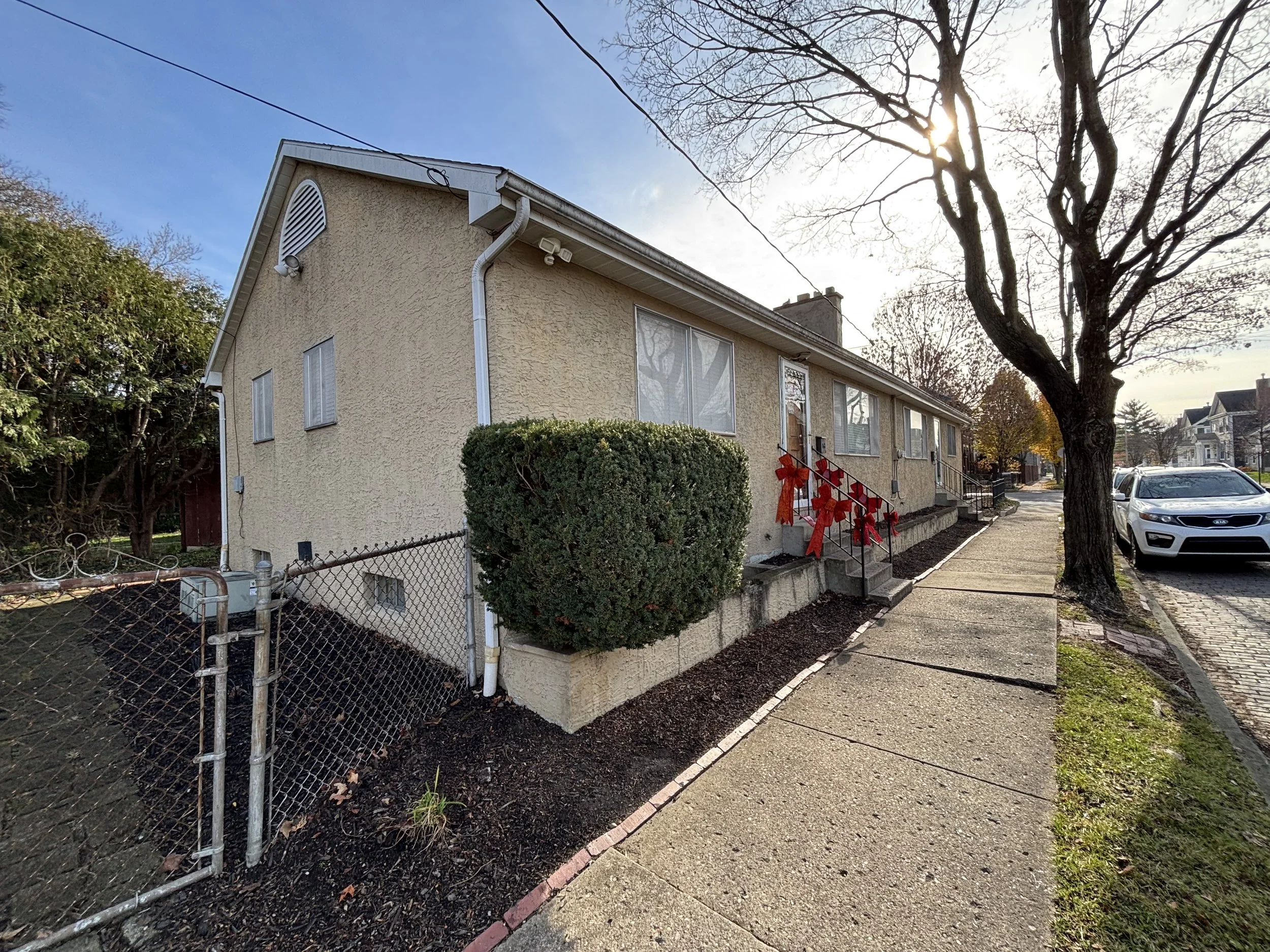 A beige residential house with a small front yard decorated with red bows on the front porch stairs, a large tree with no leaves, and a sidewalk along the street with parked cars, under a partly cloudy sky with the sun shining.