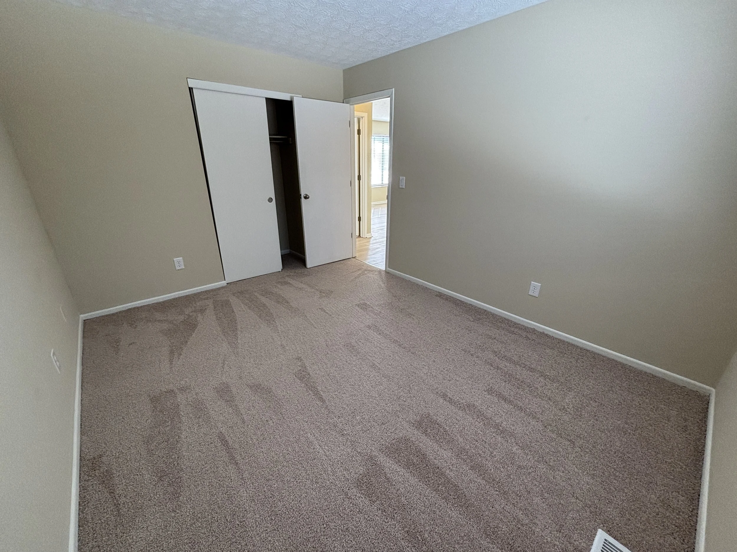 Empty bedroom with beige walls, carpeted floor, open closet door, and a doorway leading to another room.