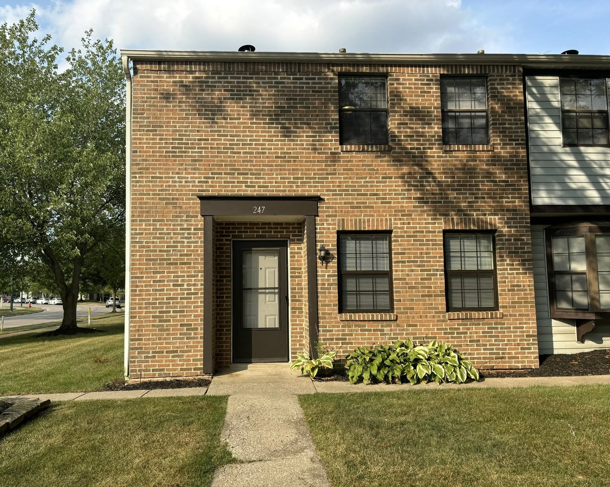 Front view of a two-story brick apartment building with a black door, four black-framed windows, and a small garden with green plants, under a cloudy sky.