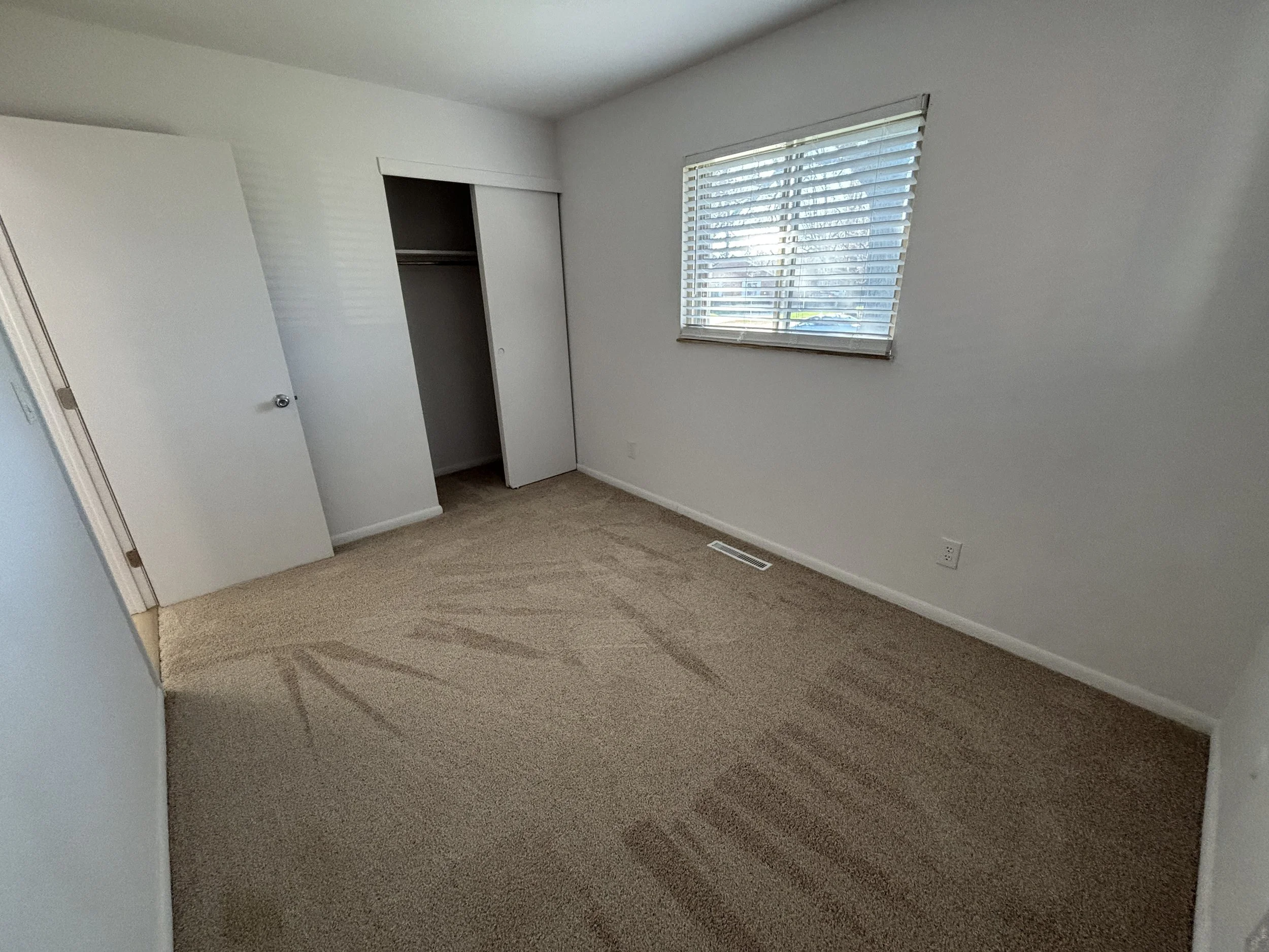 Empty bedroom with beige carpet, white walls, a window with blinds, a closed closet door, and an open closet door.