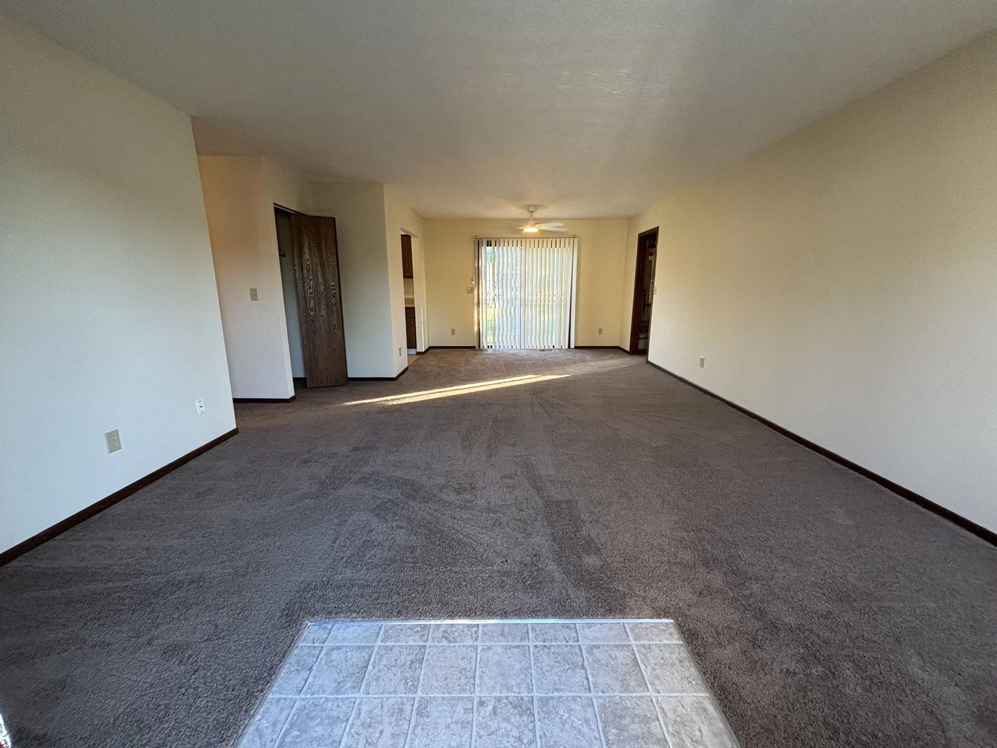 Empty living room with brown carpet, white walls, and a tile area near the entrance. There are sliding glass doors with vertical blinds leading to an outside area, and a ceiling fan in the middle of the room.