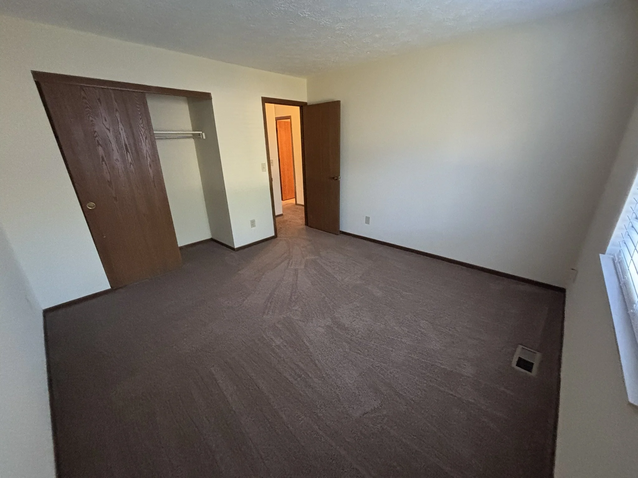 Empty bedroom with beige walls, brown carpet, a closet with wooden sliding doors, and a window with blinds.