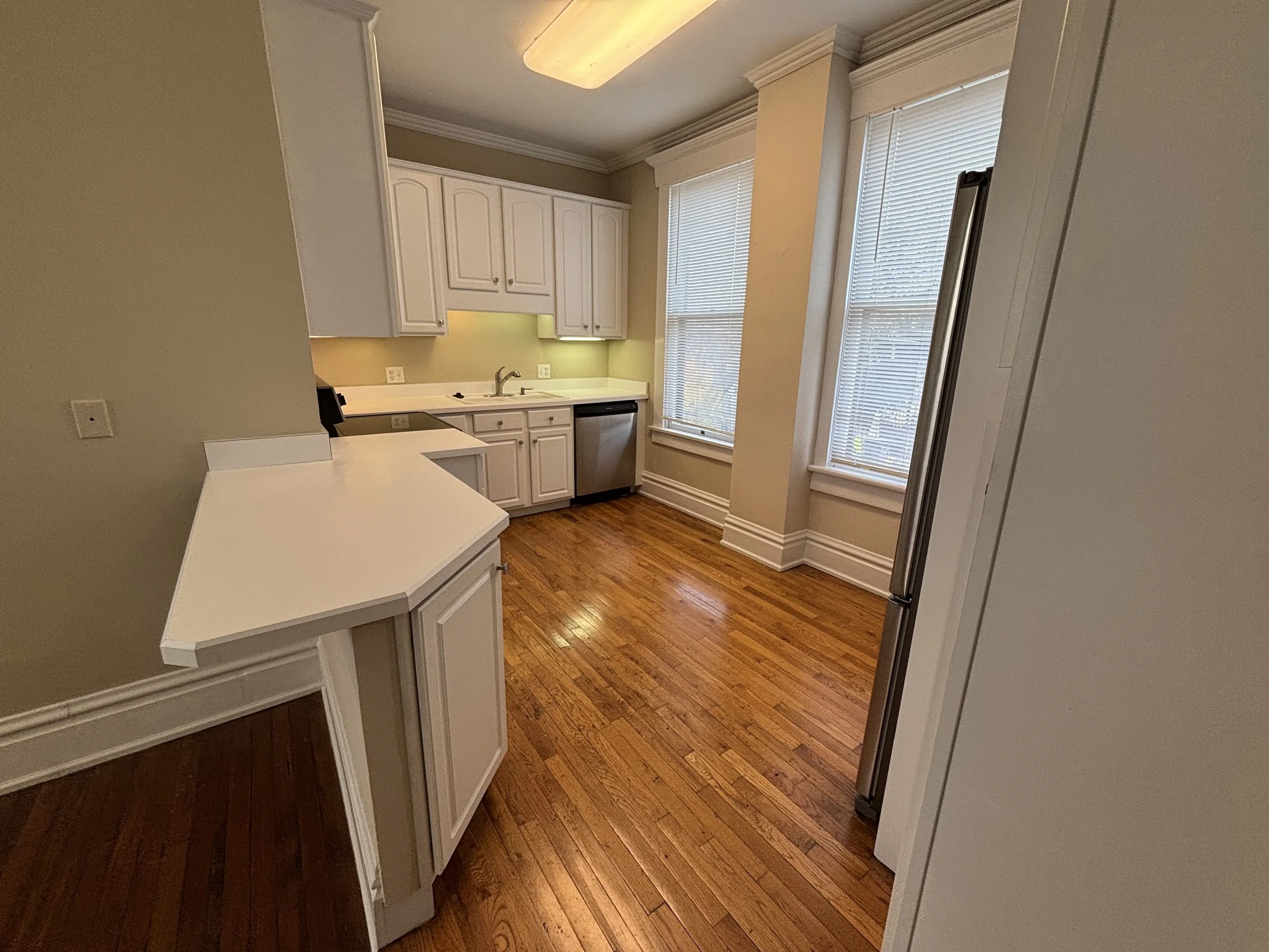 Empty kitchen with white cabinets, wooden floor, two large windows with blinds, and stainless steel appliances.