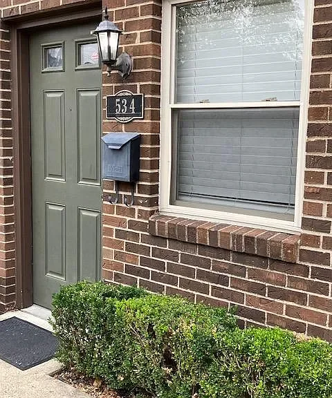The image shows a green front door with a black house number plaque reading 534, a black mailbox, a black outdoor light fixture, a window with white blinds, and a trimmed hedge in front of a brick wall.