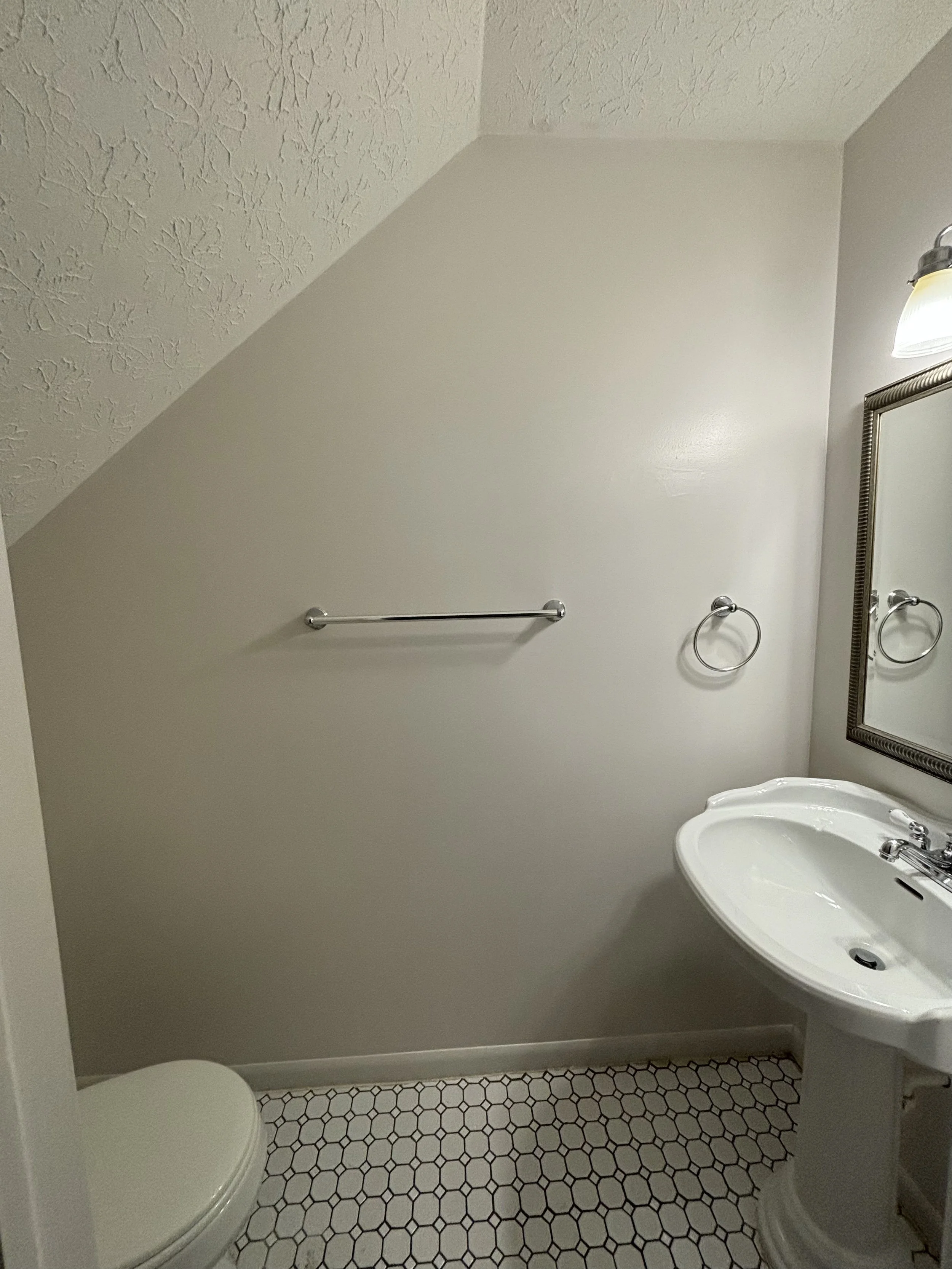 A small bathroom with a slanted ceiling, a white toilet on the left, a white pedestal sink on the right, a mirror above the sink, a wall-mounted light fixture, and a towel ring next to the mirror.