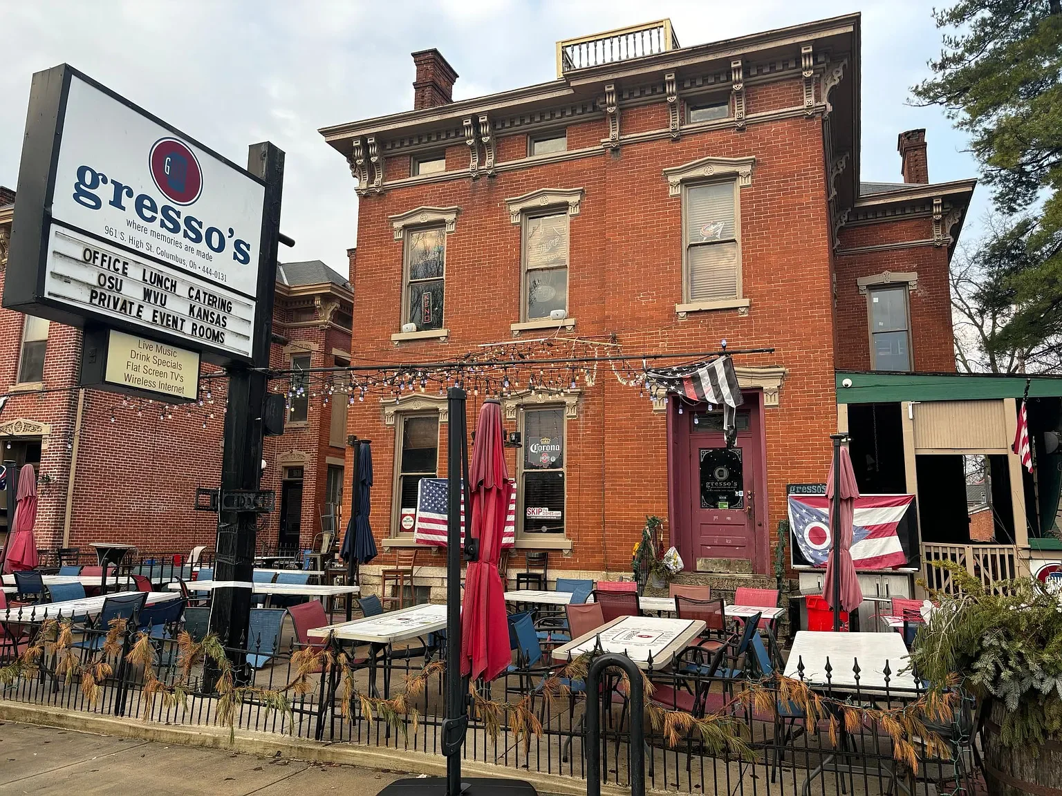 Exterior of a brick building with a restaurant or bar named Gresso's, with outdoor seating area decorated with string lights and American flags, and a sign listing offerings like office space, lunch, catering, and private event rooms.