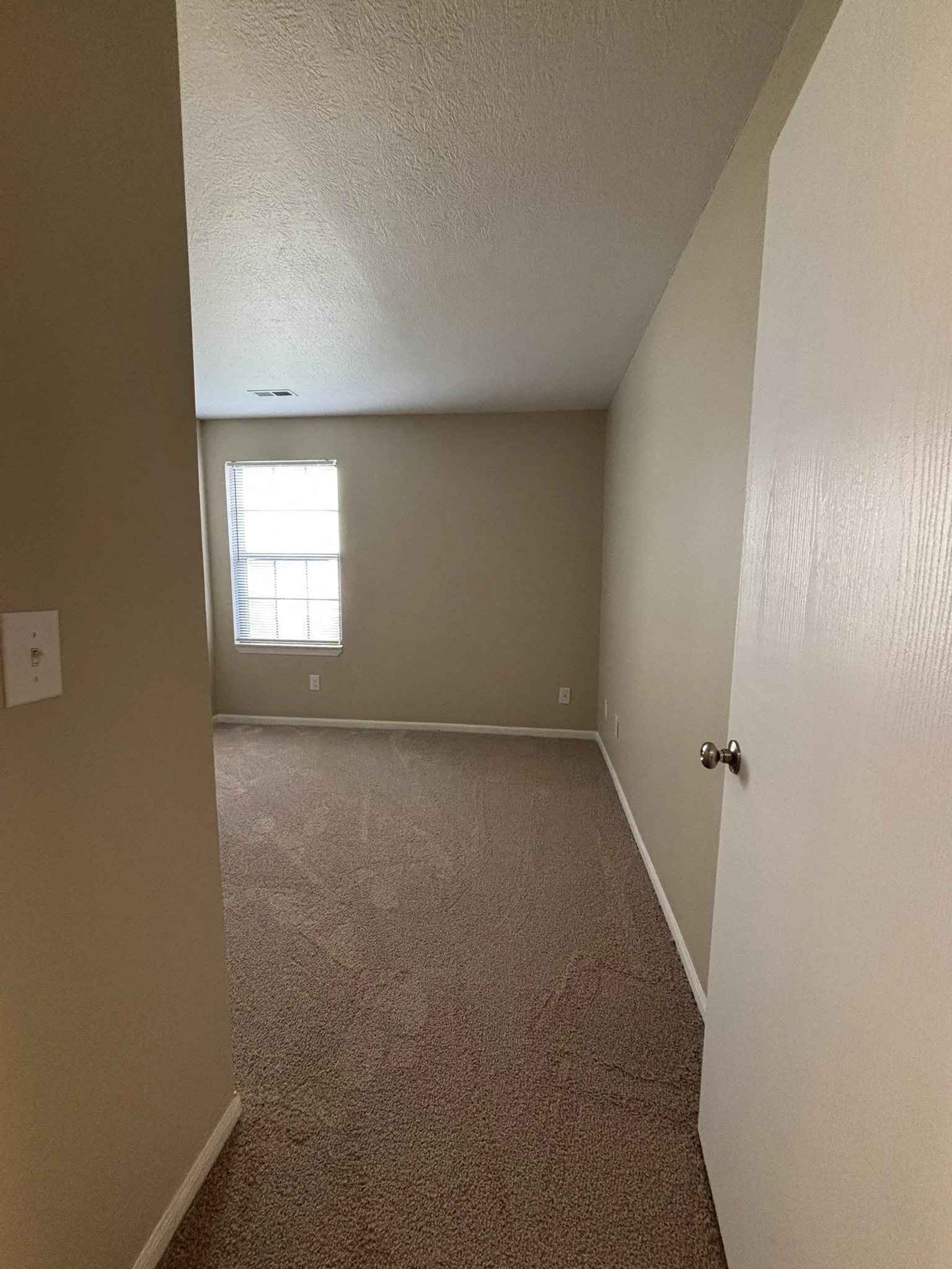 An empty room with beige walls, a window with blinds, a carpeted floor, and a partially open white door.