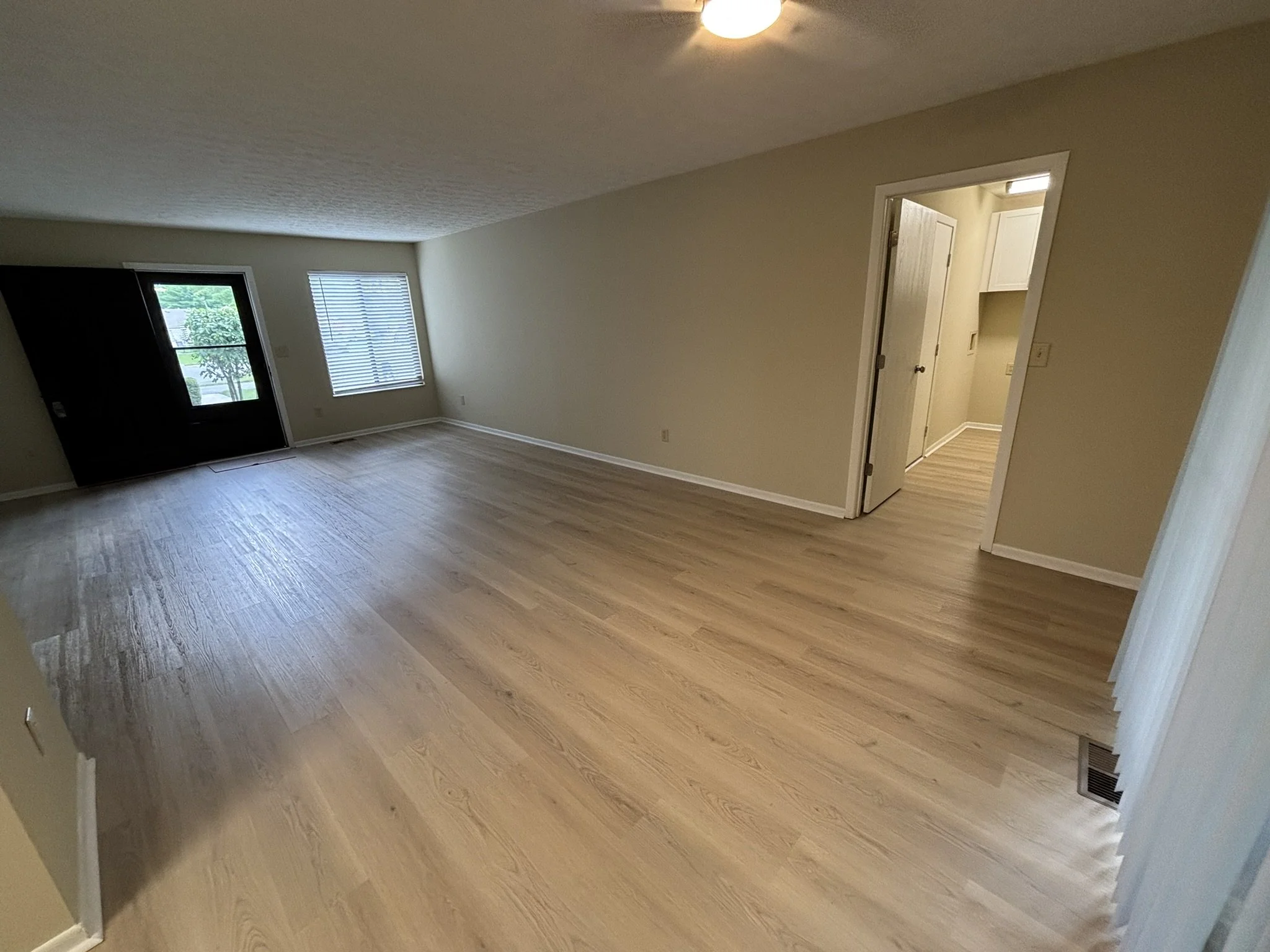Empty living room with light-colored wooden flooring, beige walls, two windows with blinds, a door leading outside, and a small doorway to another room.
