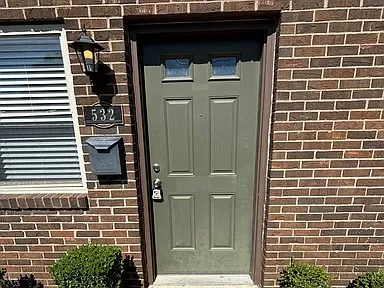 A green front door with four rectangular panels, a silver handle, and a deadbolt. The door is set in a brick wall with a small window and a black lantern-style light fixture to the left. There is a black mailbox and house number 532 on the wall. Two 