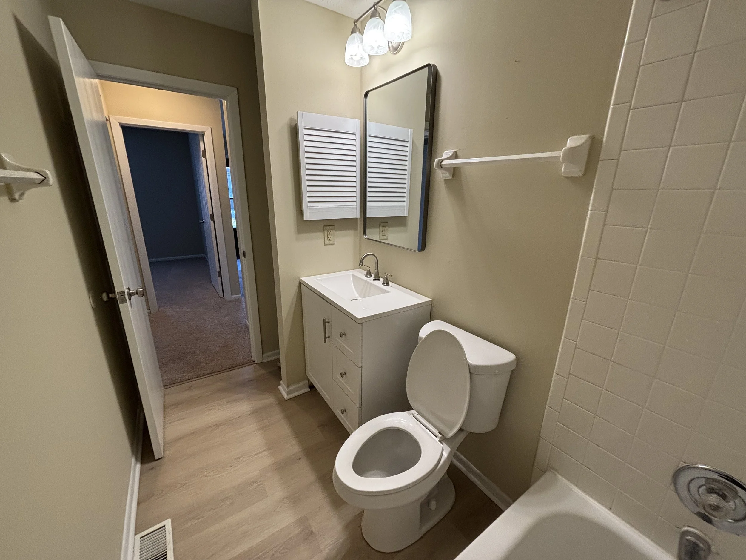 Small bathroom with a white toilet, small white vanity with a sink, mirror, wall-mounted lights, towel rack, beige walls, and part of a shower stall with white tiles.
