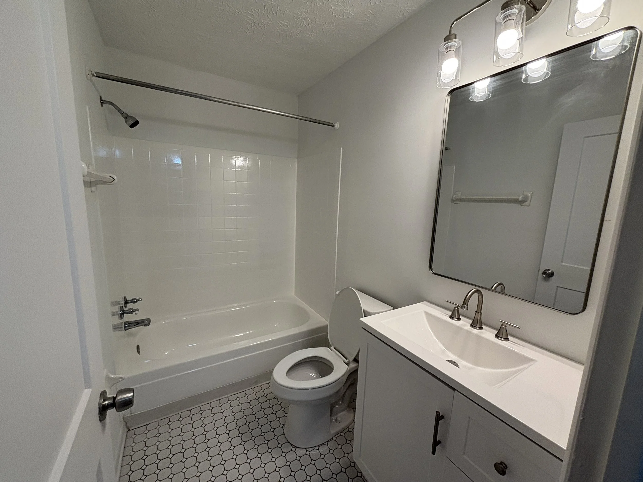 A small bathroom with a bathtub and shower, a toilet, a white vanity with a sink, a mirror, and a light fixture with multiple bulbs. The floor has hexagonal tiles.
