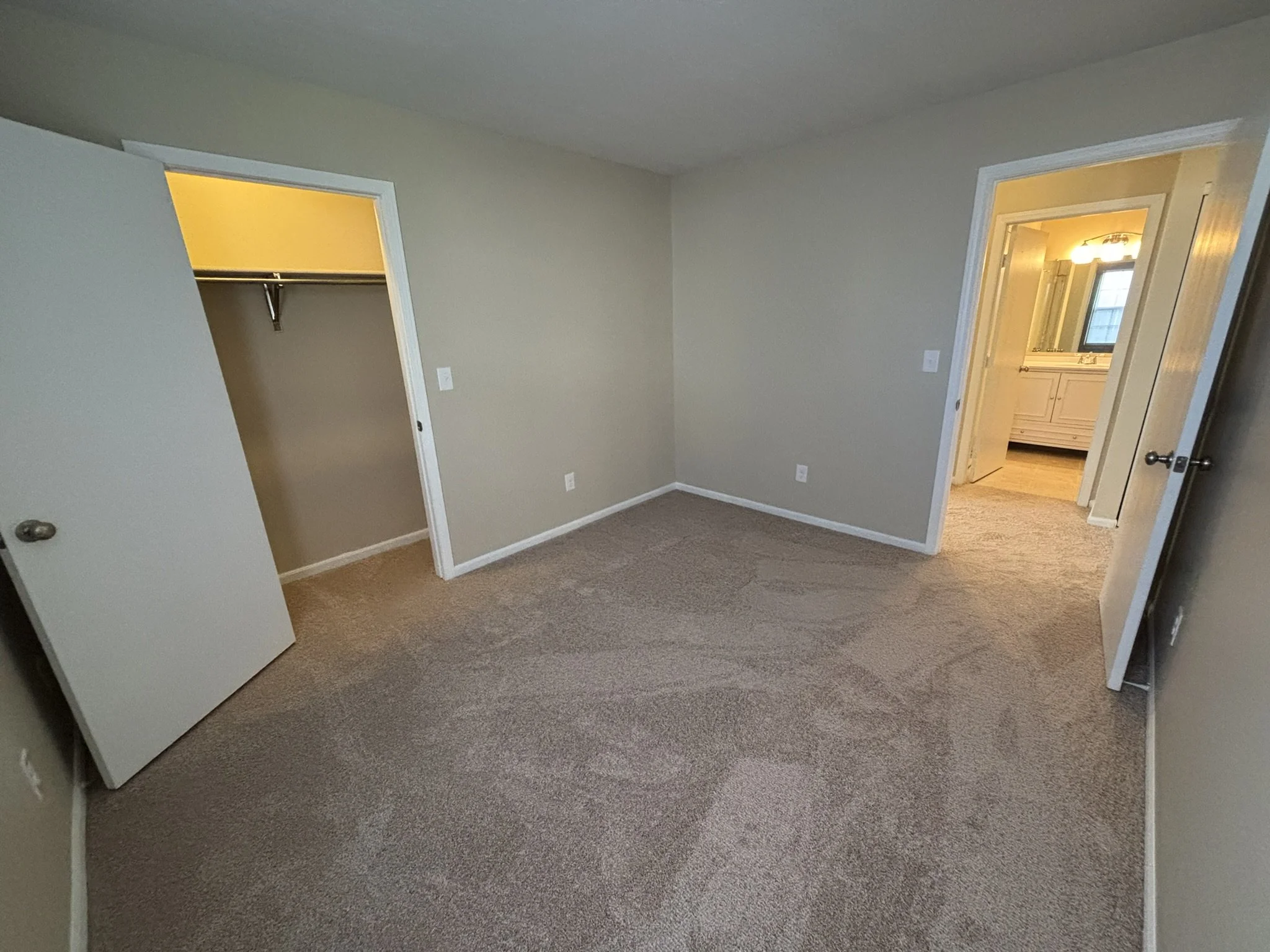 Empty bedroom with beige walls and carpet, open closet with yellow light, and an open door leading to a bathroom with white vanity and mirror.