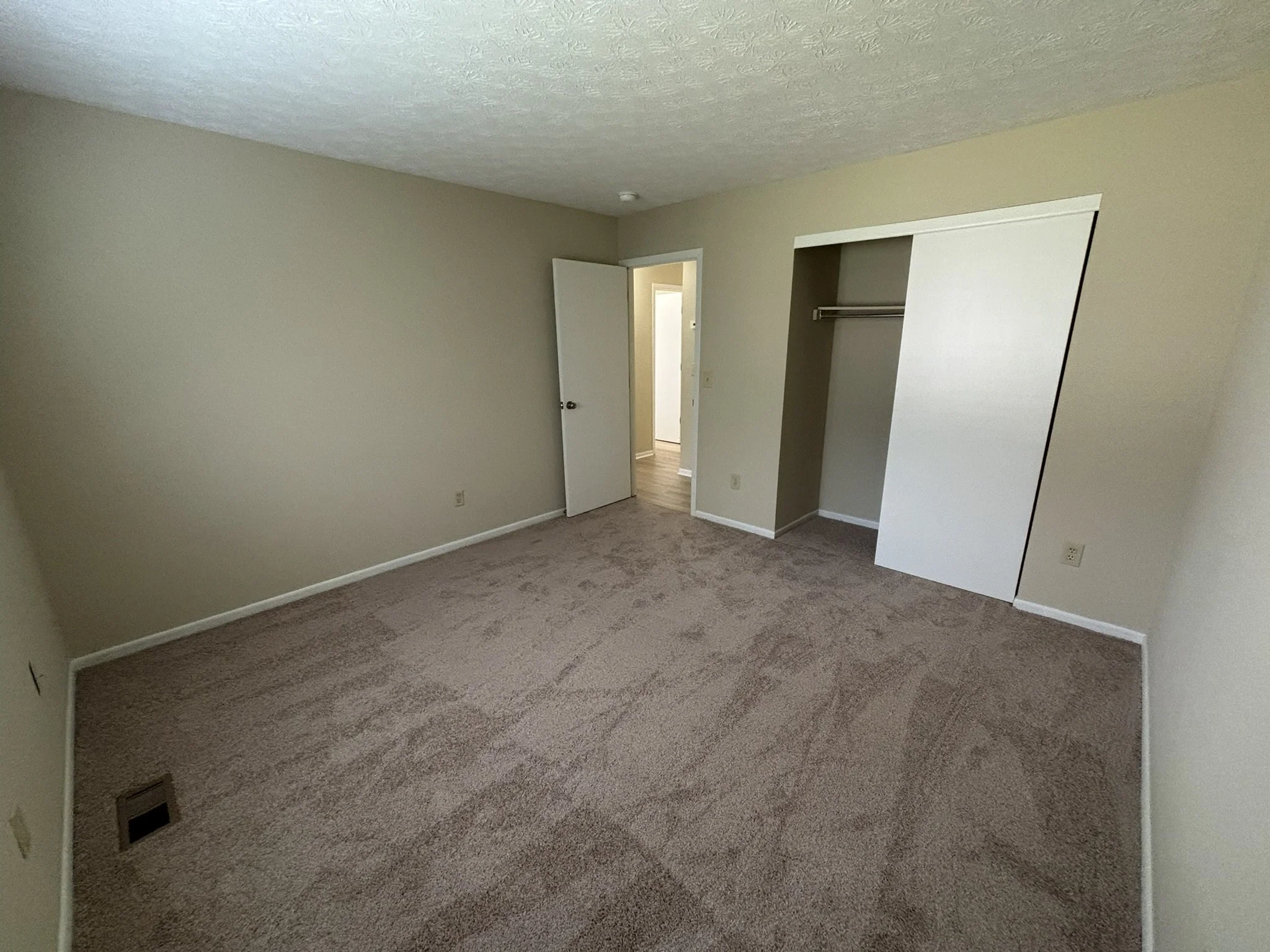 Empty bedroom with beige carpet, white walls, a closet with a sliding door, and a doorway leading to a hallway.