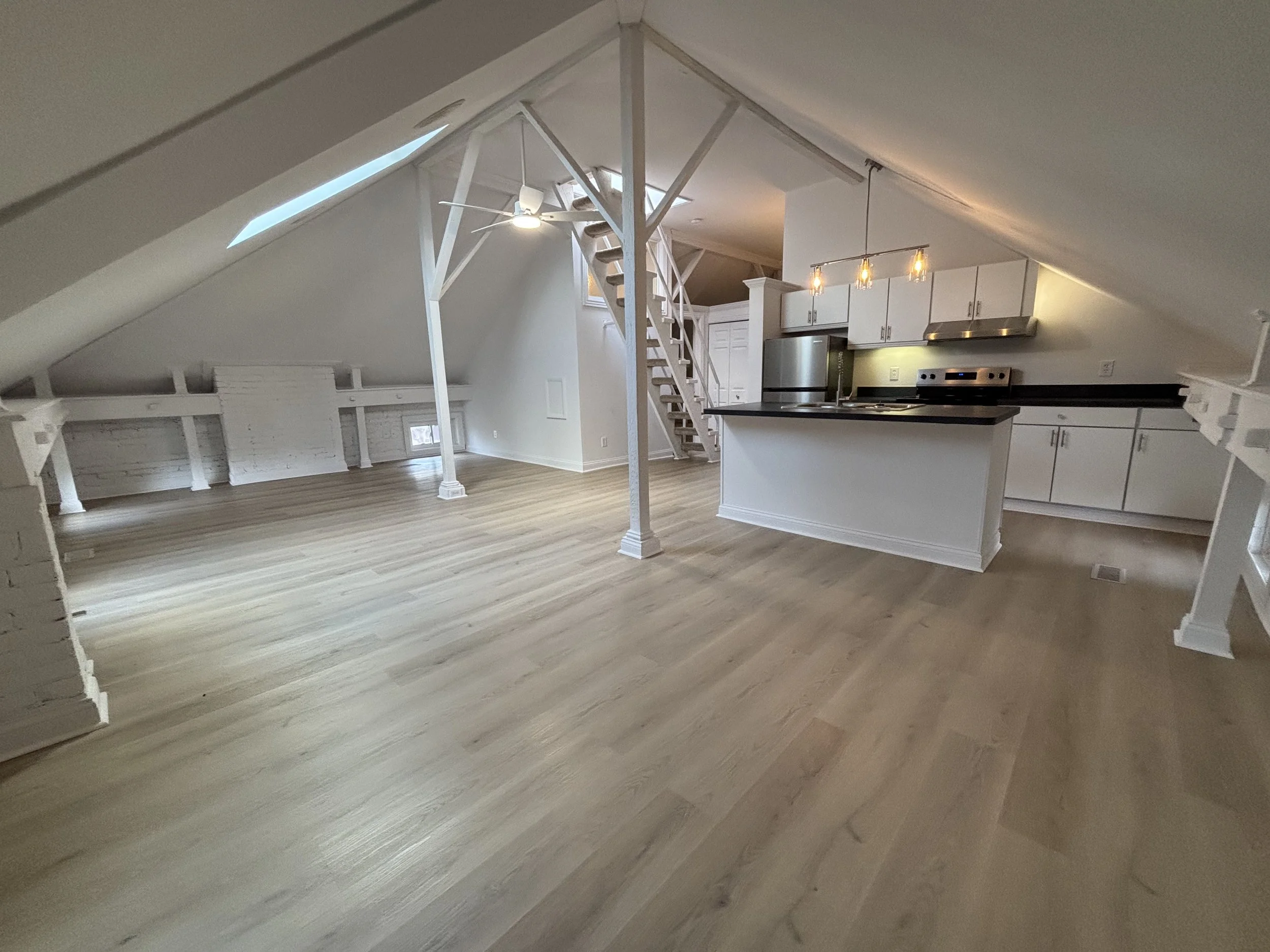 Open-concept attic apartment with hardwood floors, white walls, a small kitchen with white cabinets, stainless steel appliances, and a black countertop, illuminated by track lighting and a ceiling fan, with a skylight and small windows.