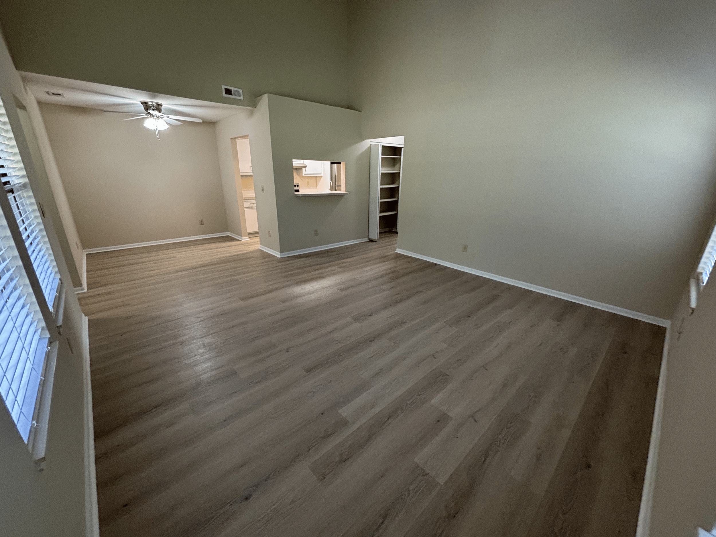Empty living room with wood laminate flooring, white walls, and multiple windows with blinds, leading into a dining area and kitchen.