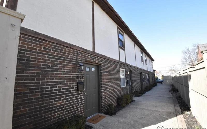 Exterior view of a row of townhouses with brick lower walls and white upper sections, a sidewalk in front, and a gray fence on the right side.