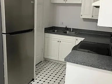 A kitchen with white cabinets, black countertops, a double sink, a refrigerator, and a tiled floor.