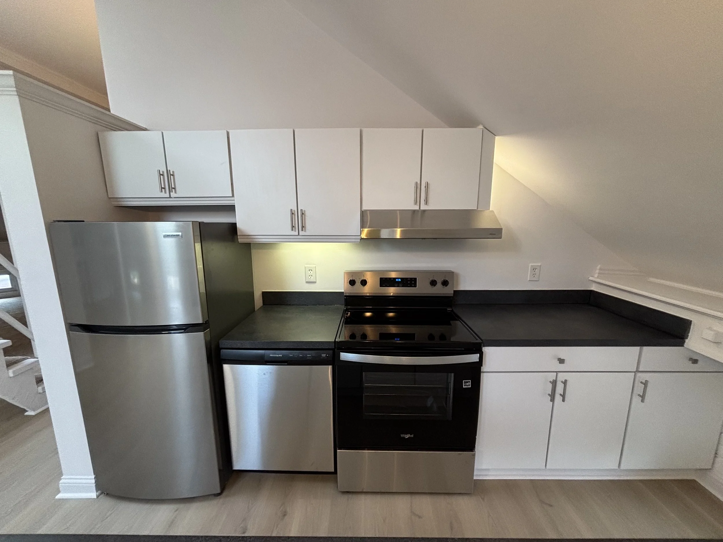 Kitchen with white cabinets, stainless steel refrigerator, black countertop, and electric stove with oven, under a sloped ceiling.