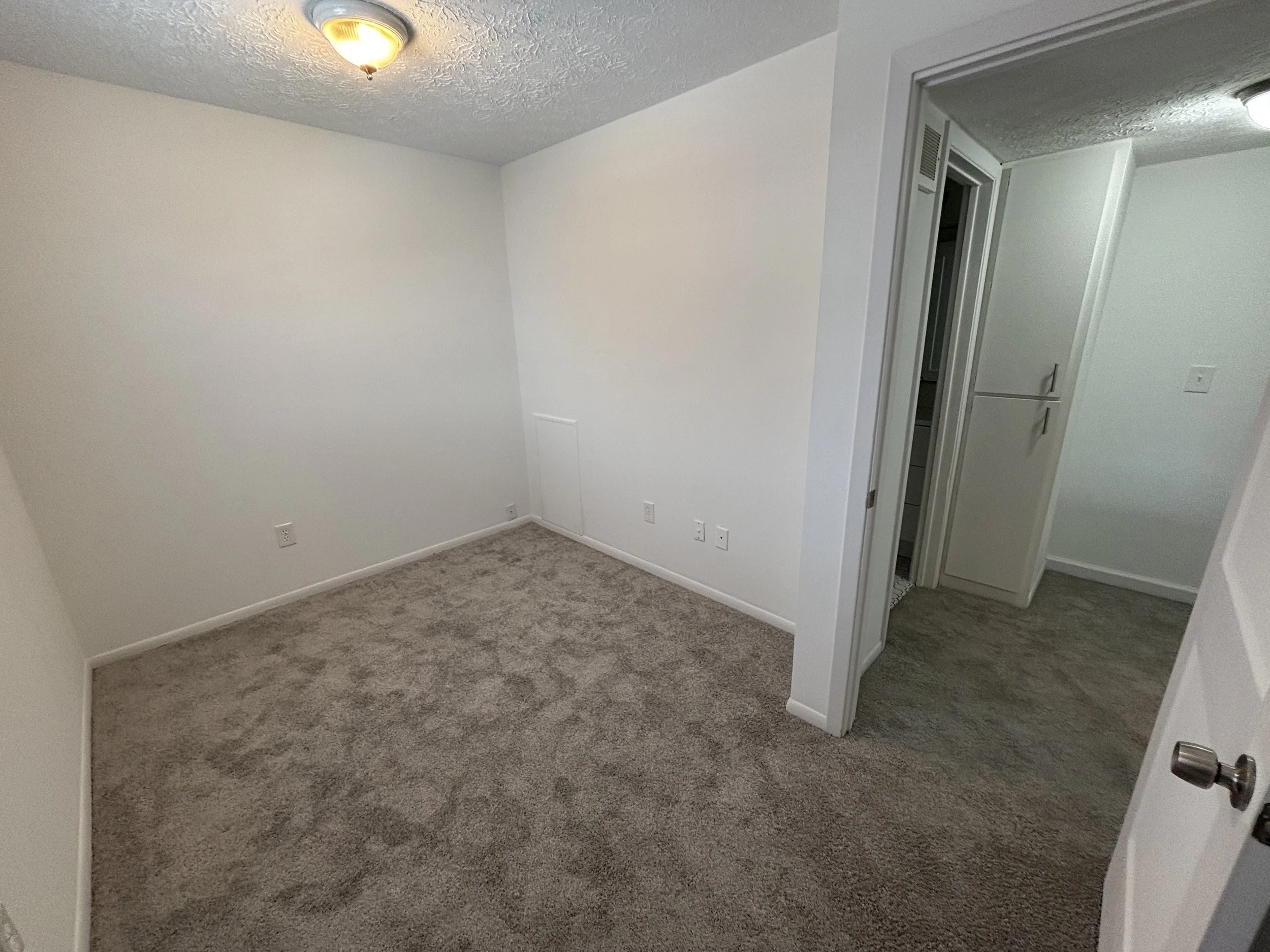 Empty white room with beige carpet flooring and a ceiling light fixture, with a doorway leading to a small closet or utility area.