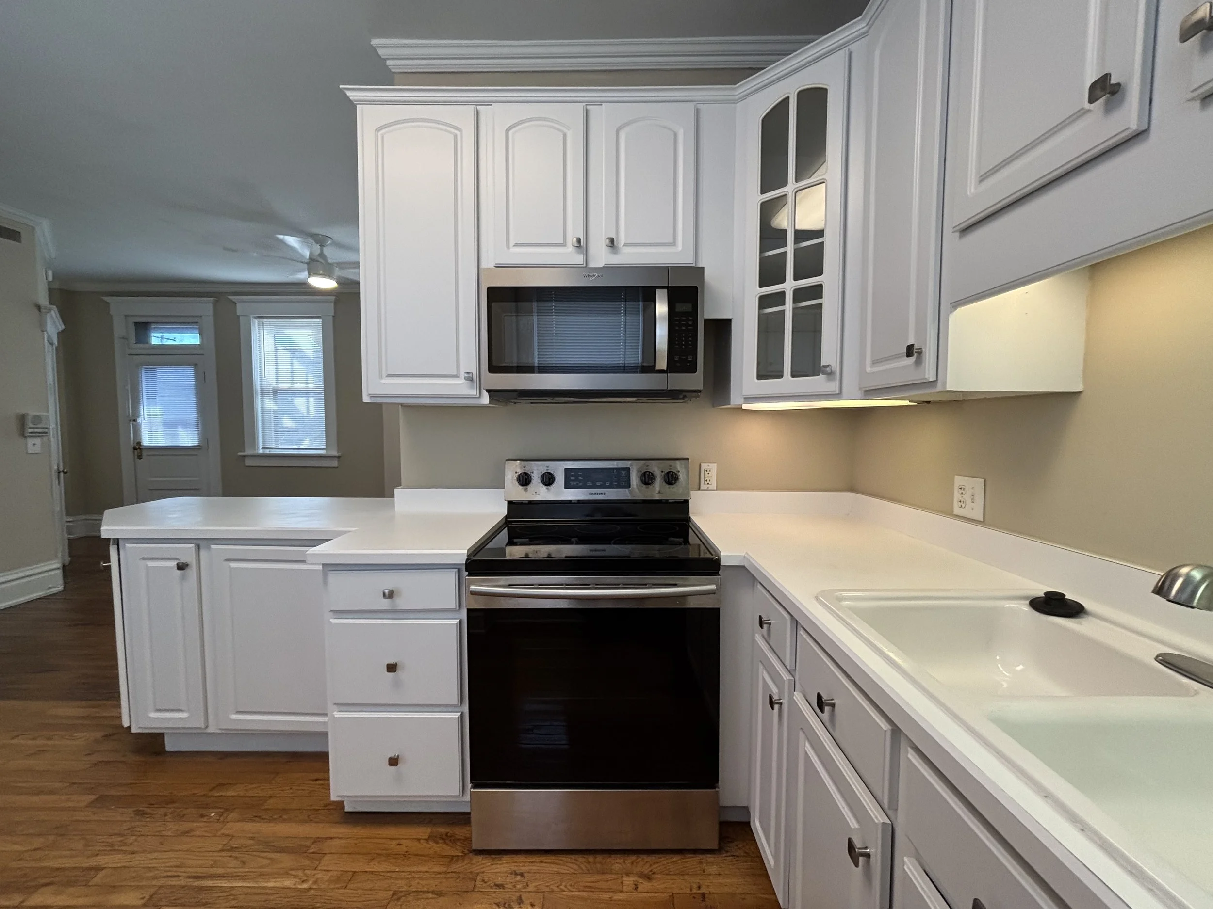 Kitchen with white cabinets, a black and stainless steel stove, a microwave above, white countertops, a double sink, hardwood floor, and windows in the background.