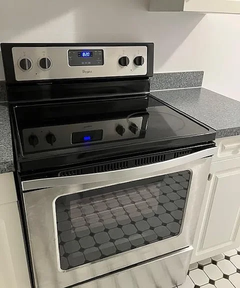 A stainless steel electric stove with a black cooktop and a digital clock, located in a kitchen with gray countertops and white cabinets.