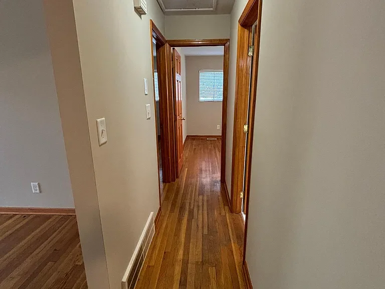 A narrow hallway with wood flooring, beige walls, wooden door frames, and a closed door at the end with a window.