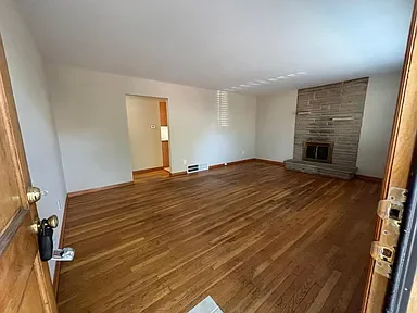 Empty living room with hardwood flooring, white walls, and a brick fireplace in the corner.