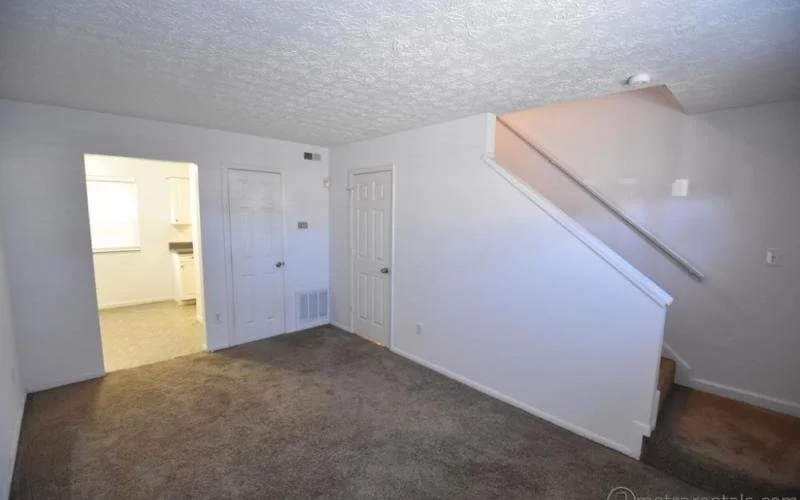 Empty living room with white walls, carpeted floor, visible staircase, and doorway leading to another room.